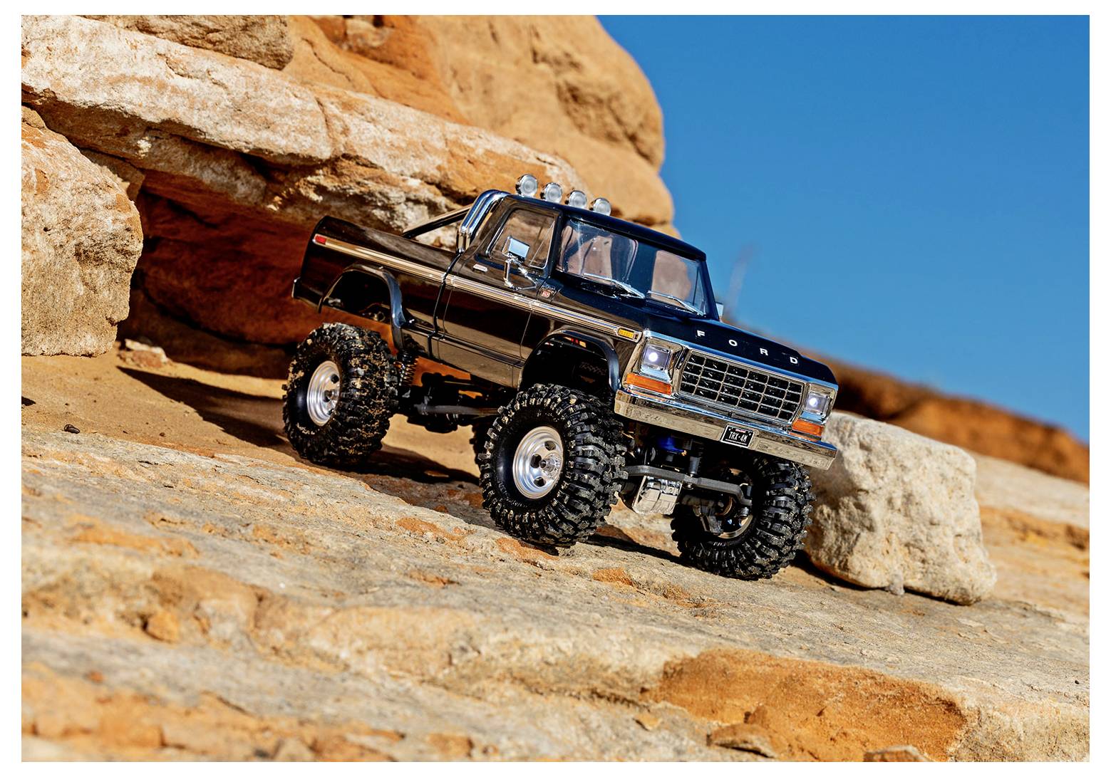 Model crnog kamiona s velikim off-road gumama stoji na stijenastom terenu ispod vedrog plavog neba.
