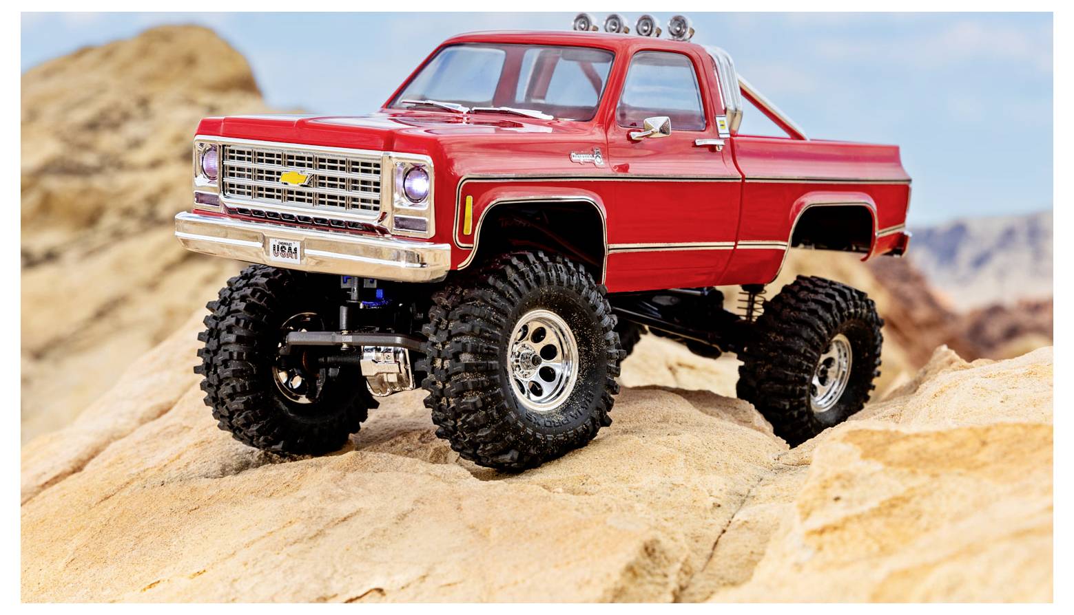 Crveni igračka monster truck s velikim gumama postavljen je na kamenovit teren, demonstrirajući svoje terenske sposobnosti. Pozadina prikazuje pustinjski krajolik.