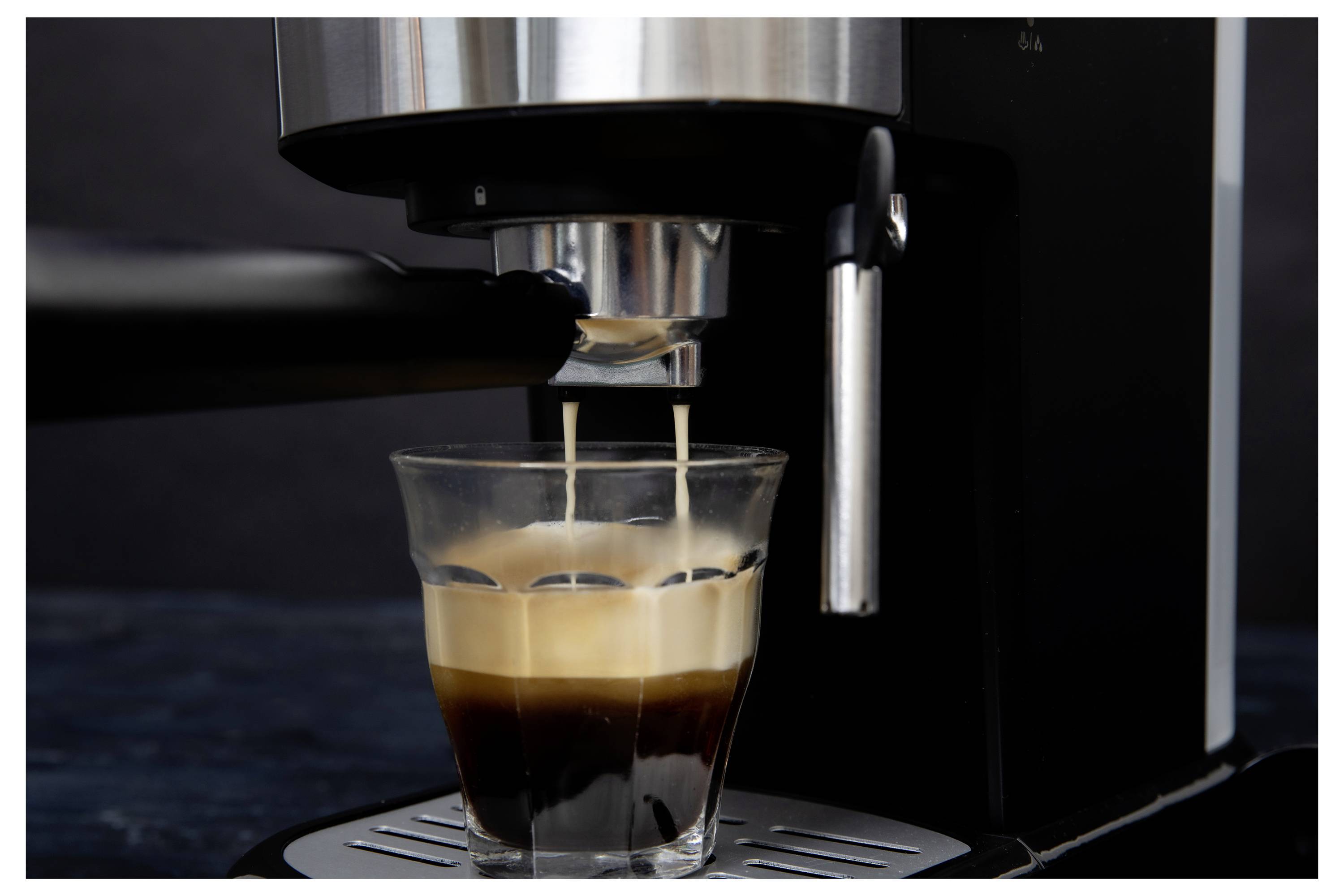 Aparat za espresso sipa vruću kavu u prozirno staklo, stvarajući slojeve kreme i tamne tekućine na tamnoj pozadini.