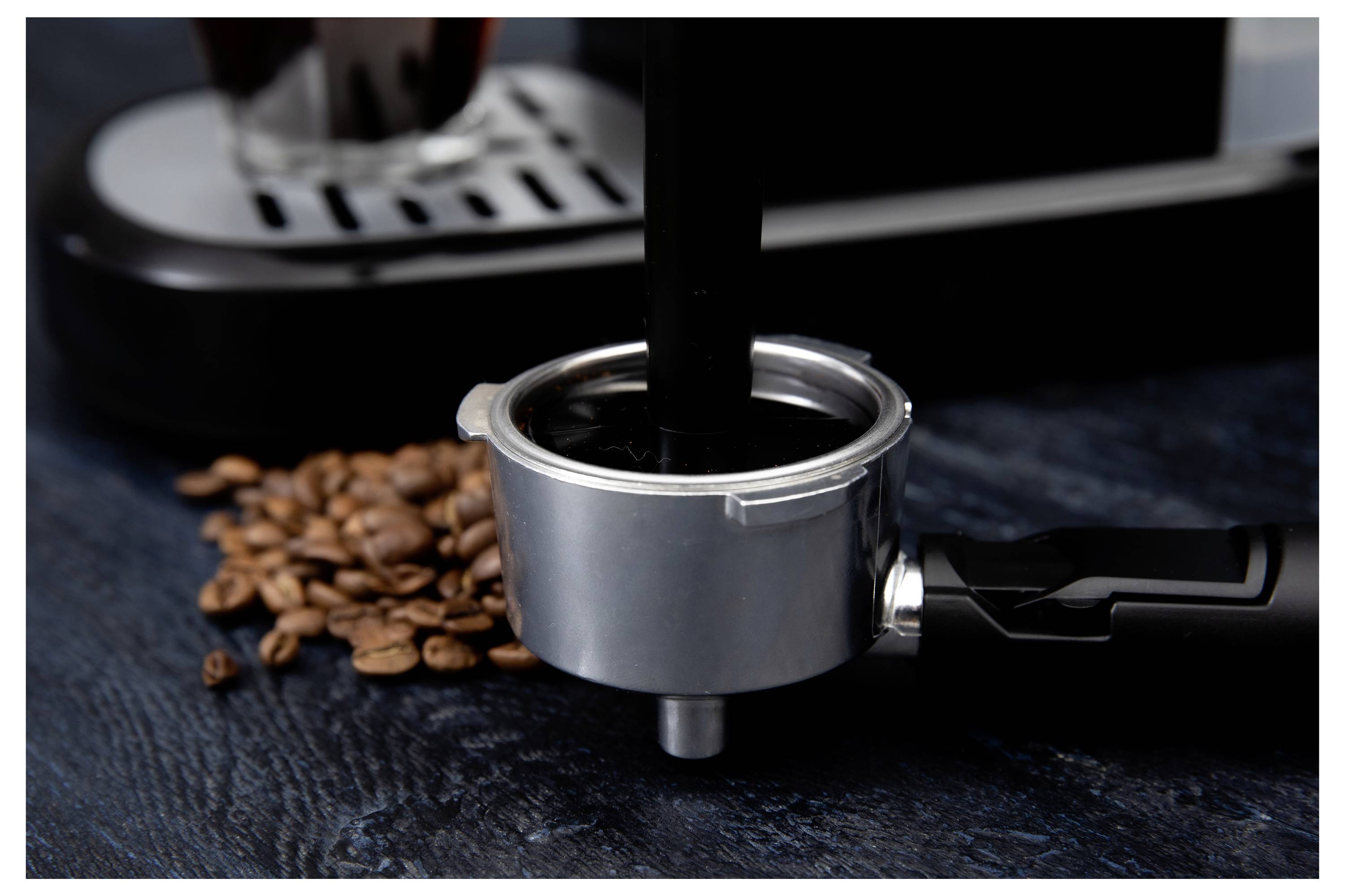 Portafilter se napunjava mljevenom kafom pokraj razasutih zrna kave. U pozadini je djelomično vidljiv aparat za espresso.