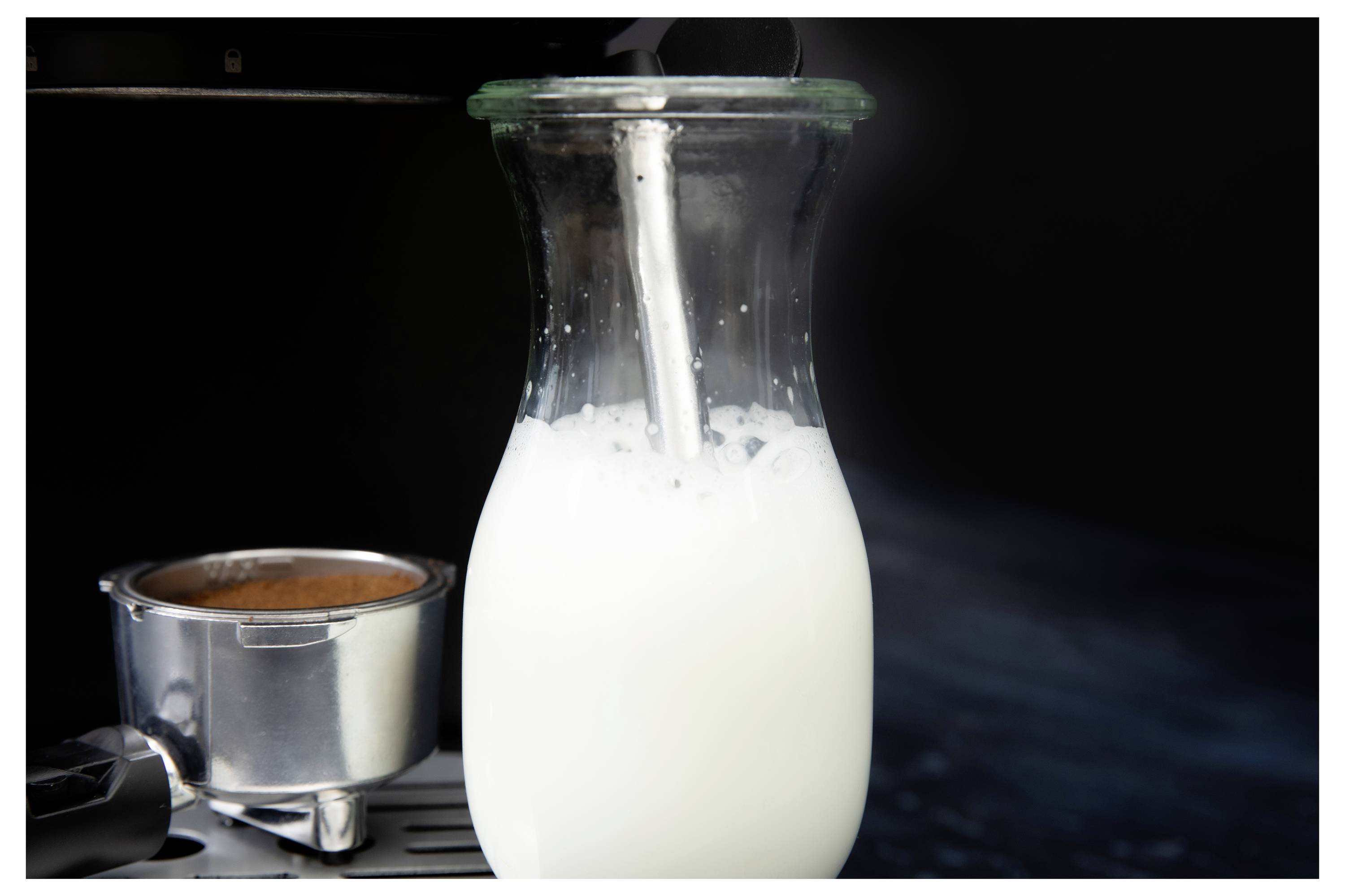 Staklena karafa napunjena mlijekom para se na aparatu za espresso, pored portafiltera s kafom, uz tamnu pozadinu.