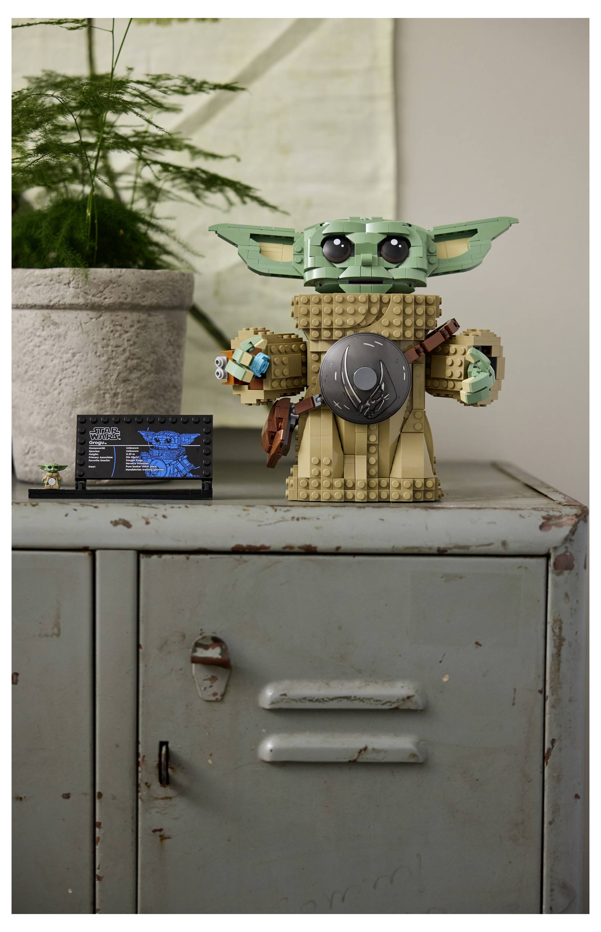Lego model koji nalikuje maloj, zelenkastoj stanoj figurici s velikim ušima stoji na metalnom ormaricu pokraj biljke u saksiji. Mala kartica s natpisom nalazi se pokraj nje.