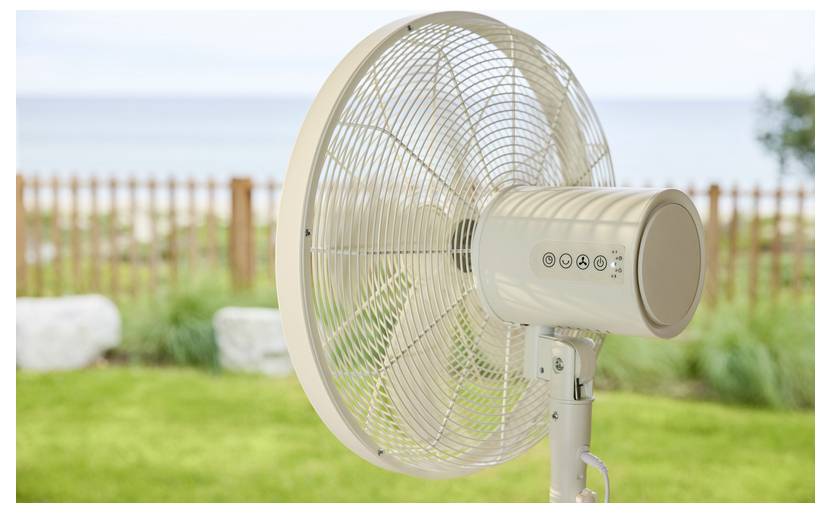 Bijeli ventilator na postolju stoji na travnatoj površini pored drvene ograde, s oceanom i nebom vidljivim u pozadini.