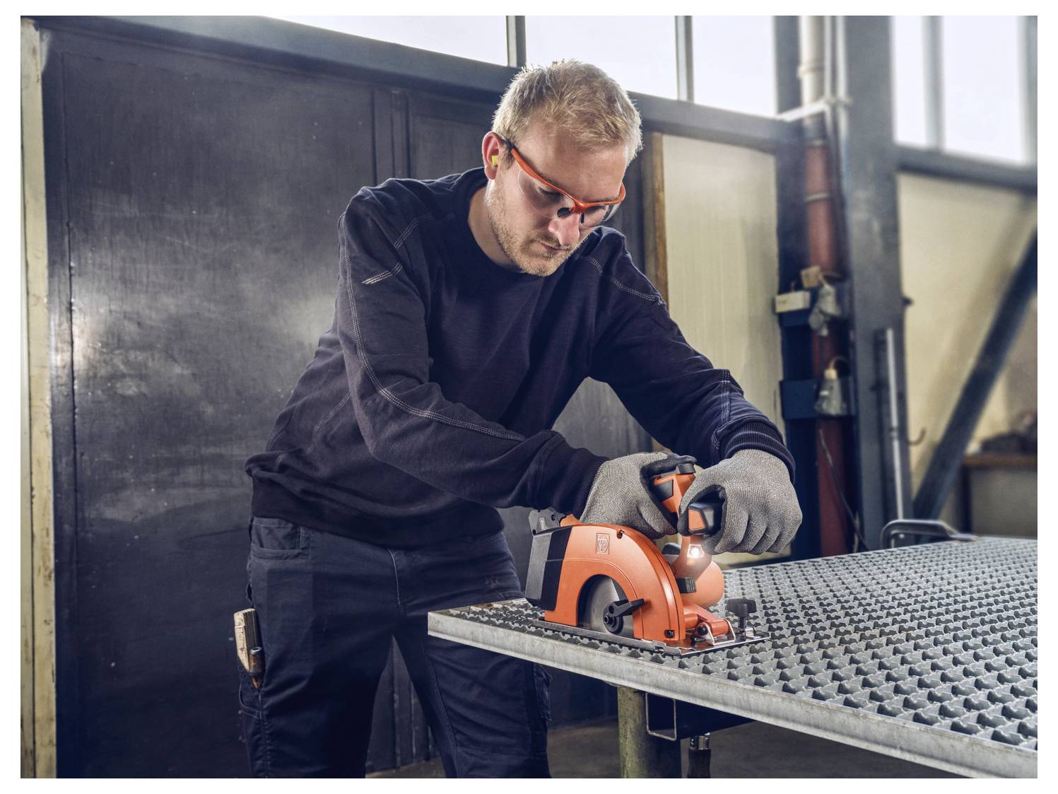 'Muškarac u zaštitnoj opremi koristi električni alat na metalnoj površini u industrijski okruženju.'