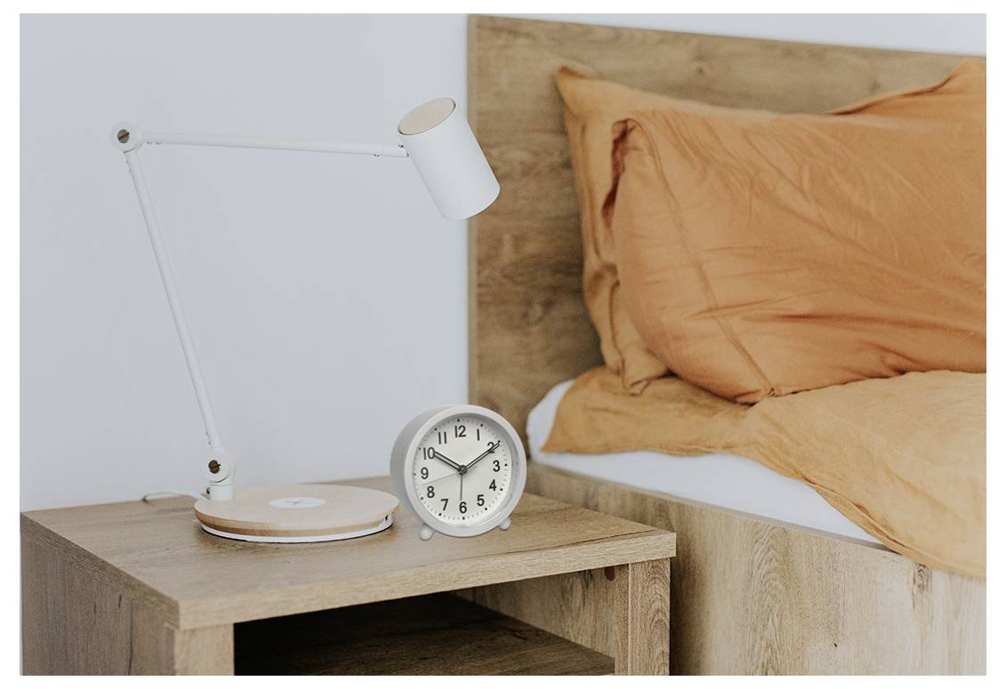 Drveni noćni stolić s bijelom lampom i okruglim srebrnim satom koji pokazuje 2:10. U pozadini je neuredjena krevet s narančastim posteljinama.