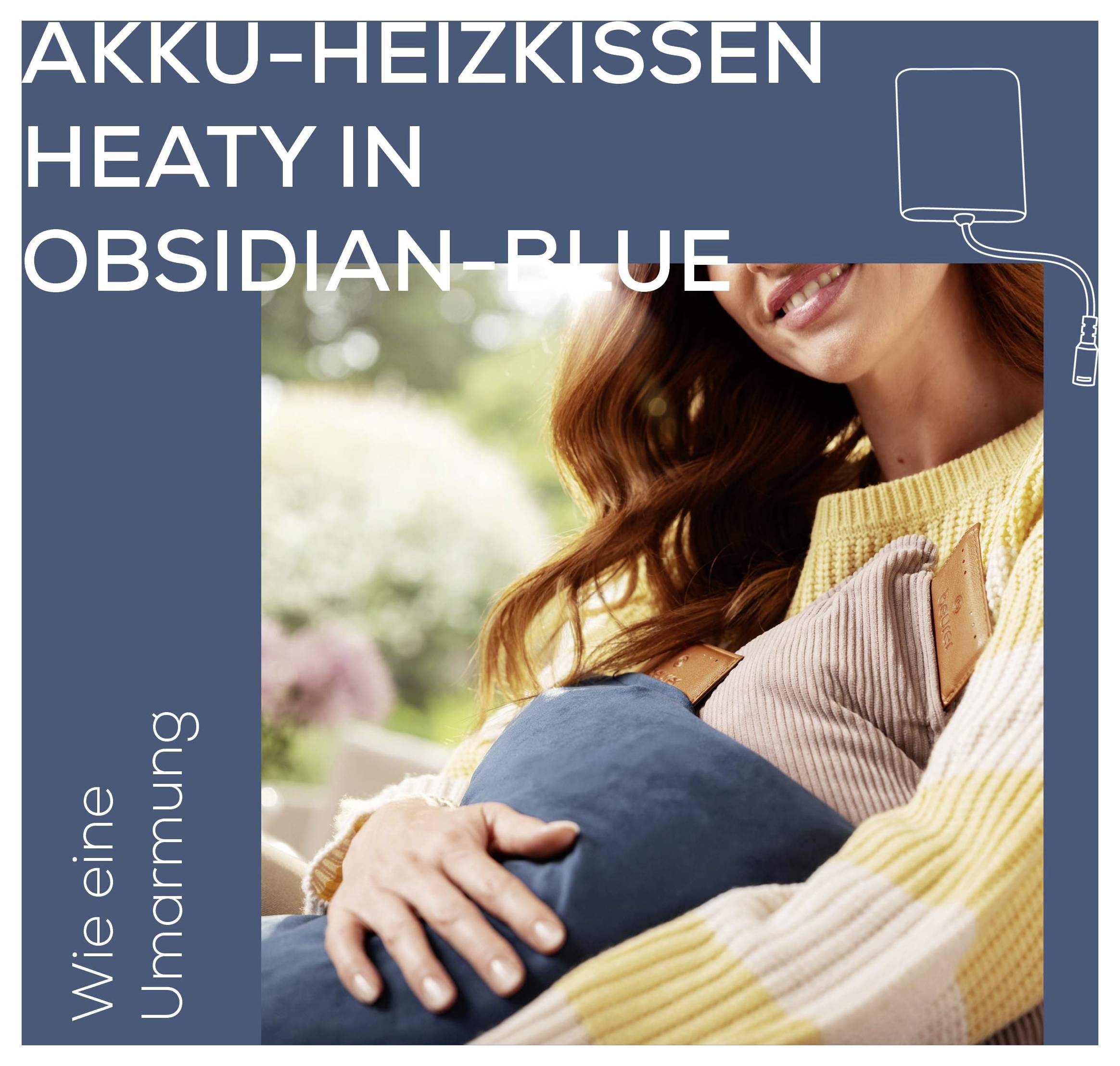 Žena, koja se smješi, drži plavi grijaći jastuk od baterije uz prsišta u zatvorenom prostoru. Tekst glasi "AKKU-HEIZKISSEN HEATY U OBSIDIJAN PLAVOJ BOJI. Kao zagrljaj."