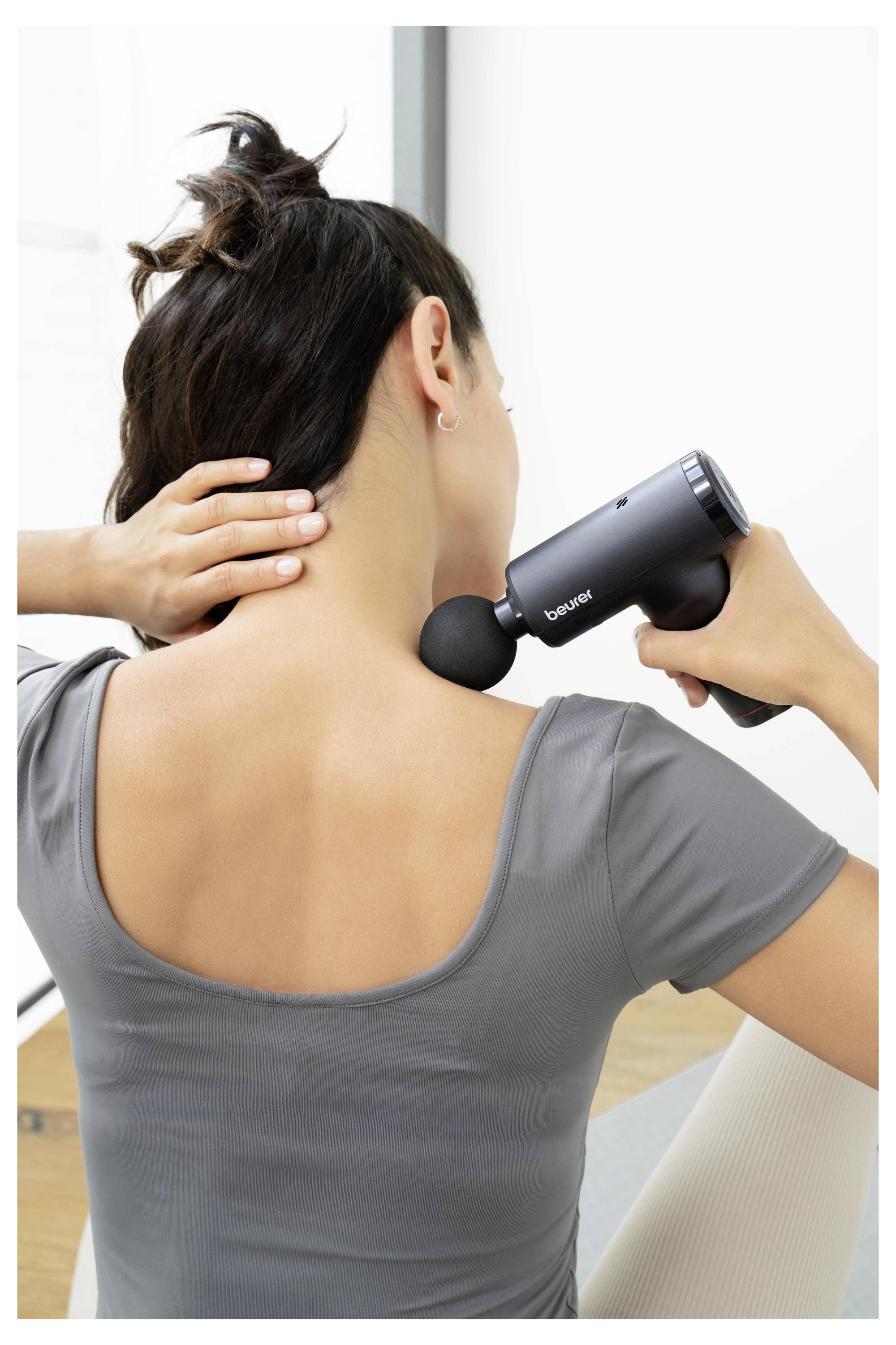 Osoba koristi crni masažni pištolj na vratu kako bi olakšala napetost mišića. Nosi sivi vrh i sjeddi u jasnoj, dobro osvjetljenoj sobi.