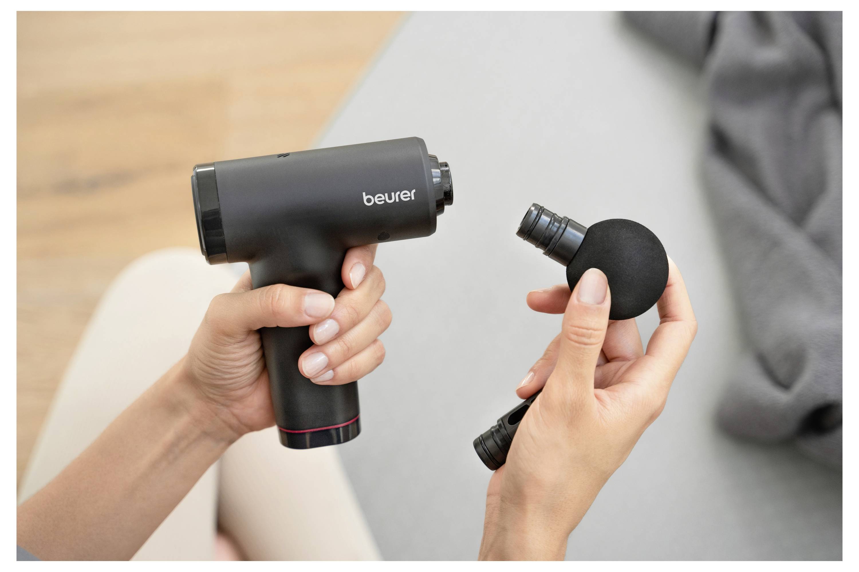 Osoba drži crni masažni pištolj u jednoj ruci, a drugom rukom pričvršćuje sfernu masažnu glavu. Blizu je siva tkanina.