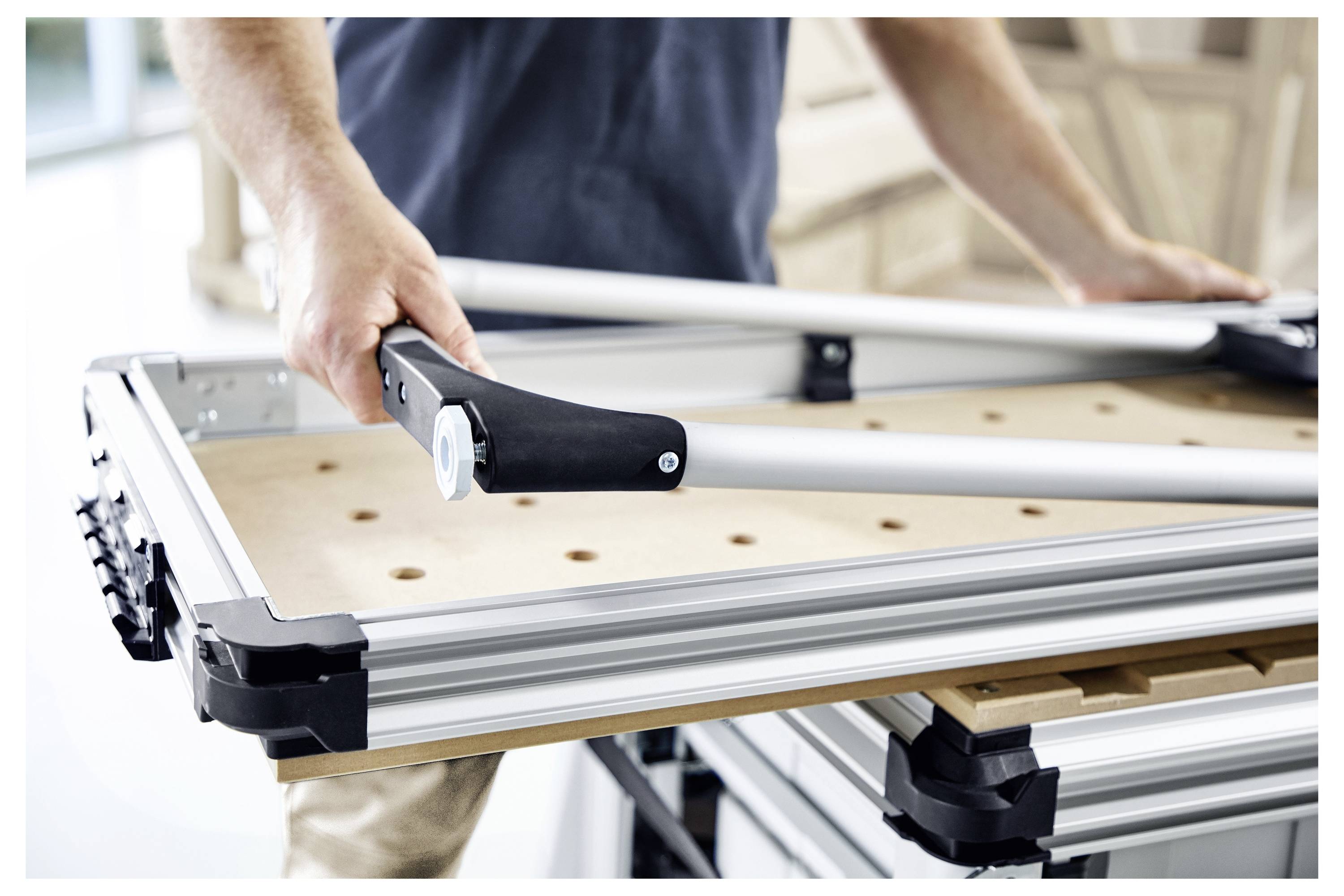 Osoba koja montira radni stol s aluminijskim okvirom i drvenom površinom, držeći металni alat u osvijetljenom radnom prostoru.