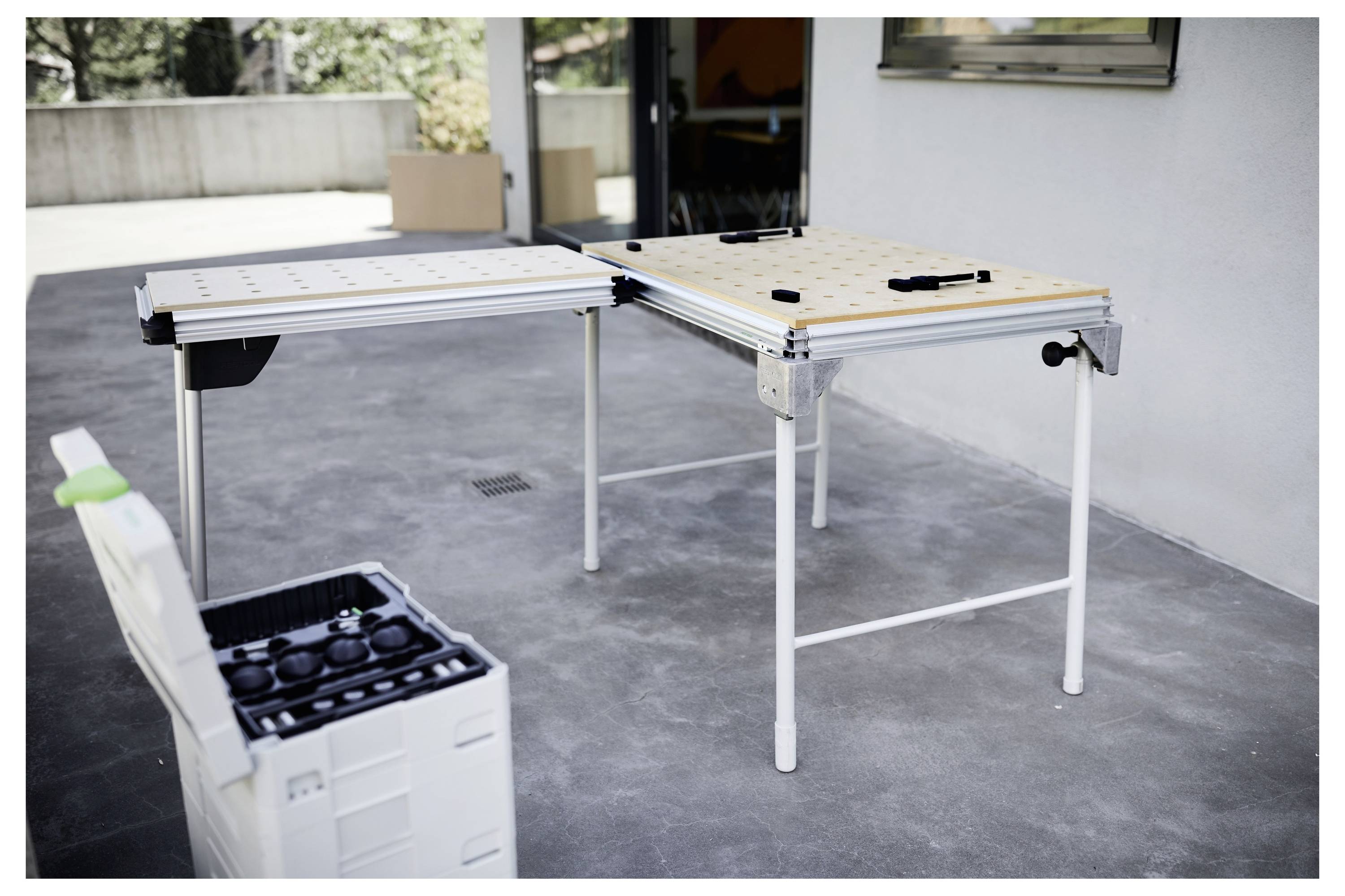 'Dva modularna radna stola sa alatima na terasi, pokraj kutije za alate koja sadrži razne spojnice. Vanjski prostor s biljkama u pozadini.'