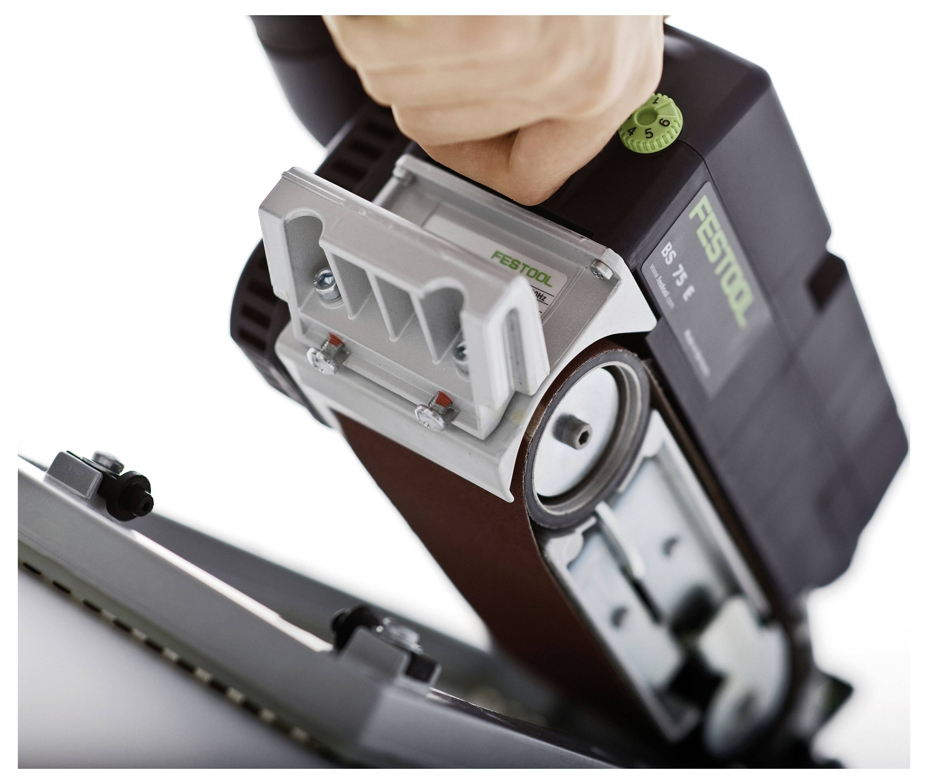 Ruka upravlja Festool trakastom brusilom, čineći površinu glatkom. Alat je nagnut, otkrivajući njegovu traku i mehanizme za podešavanje.
