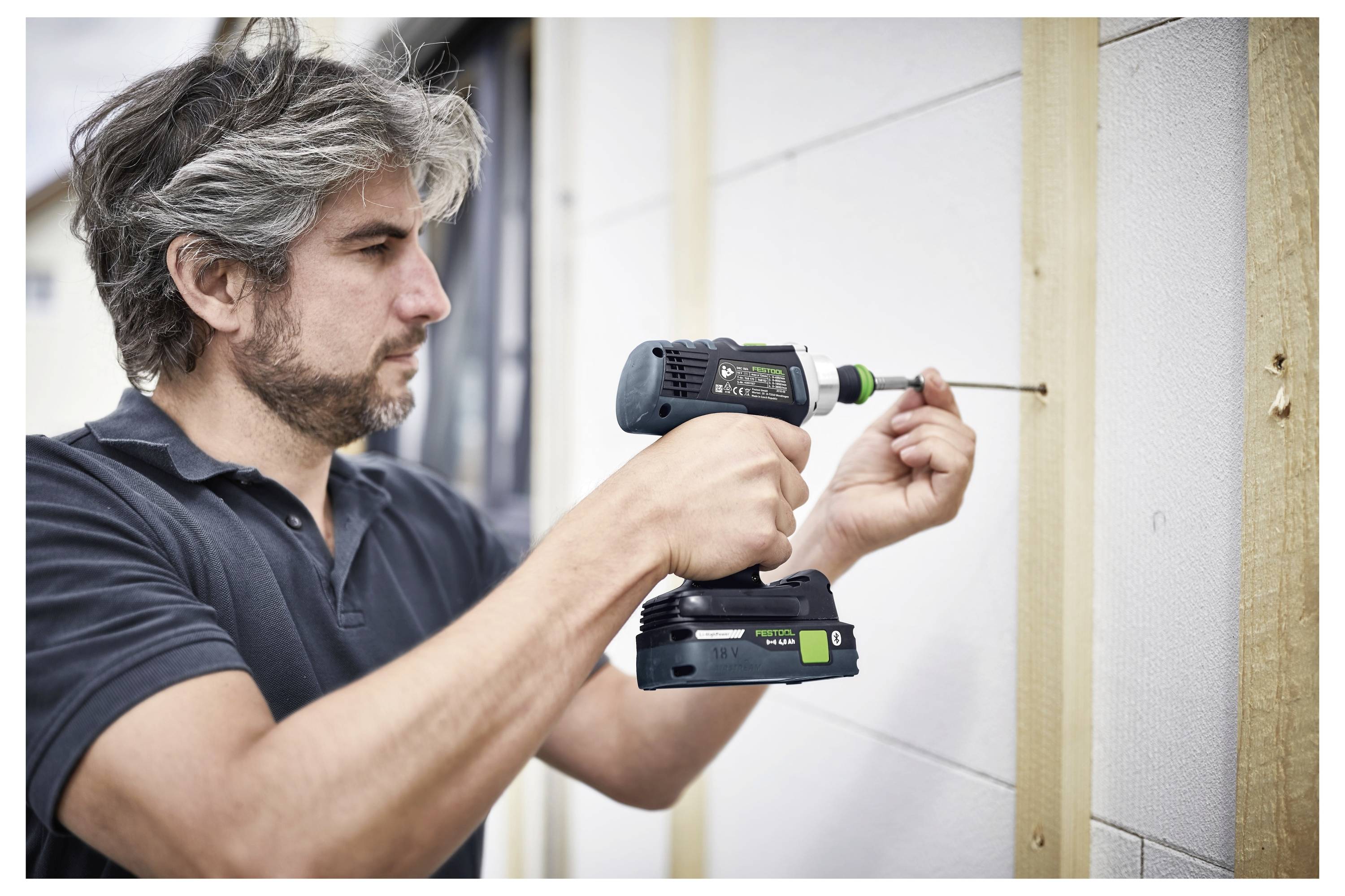Brada čovjek koristi bežičnu bušilicu da vijča drvenu ploču na bijelom zidu, intenzivno se koncentrirajući. Nosi crnu polo majicu.