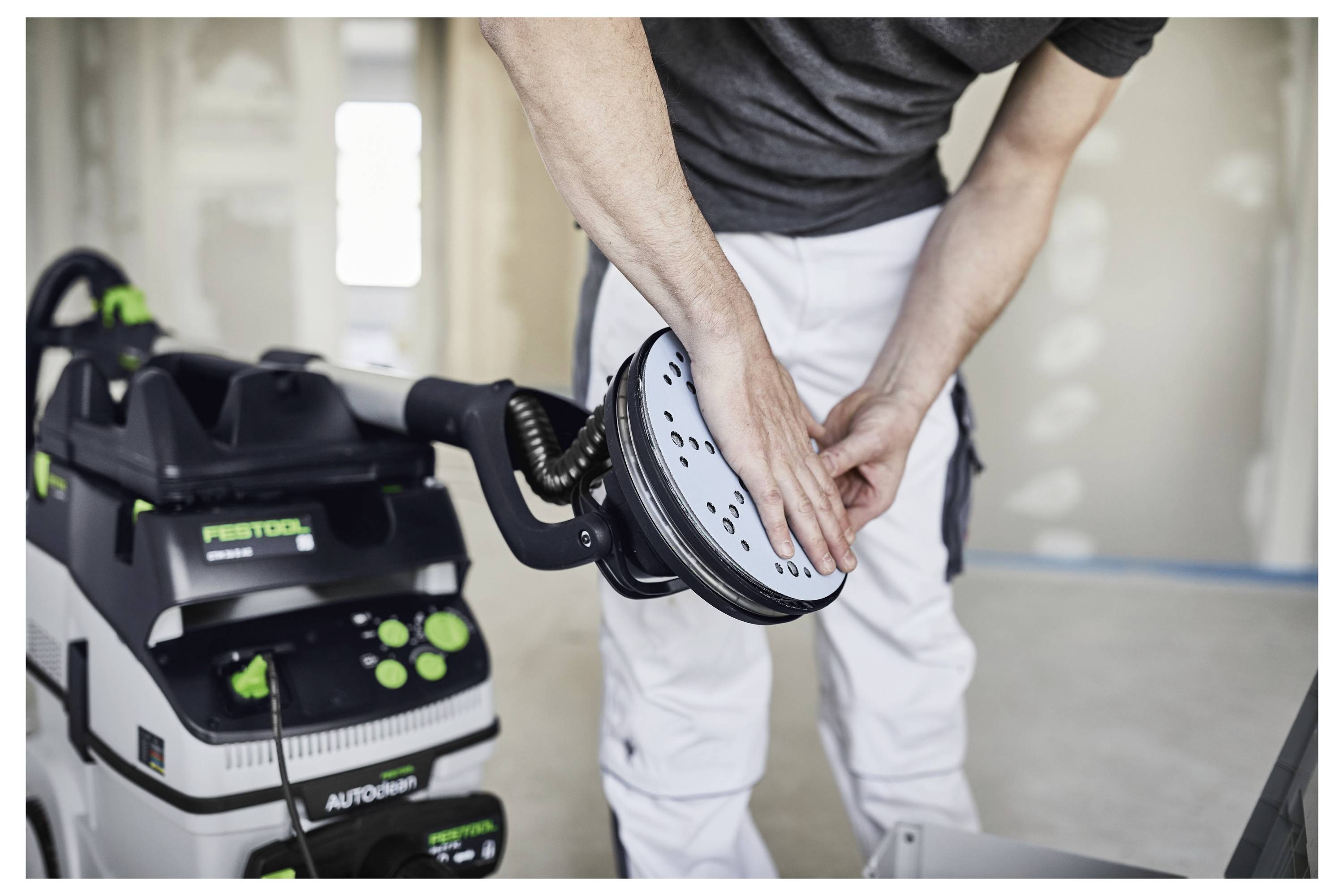 Osoba podešava ručnu brusilicu povezanu na usisavač u sobi u obnovi, noseći radne hlače i tamnu majicu.