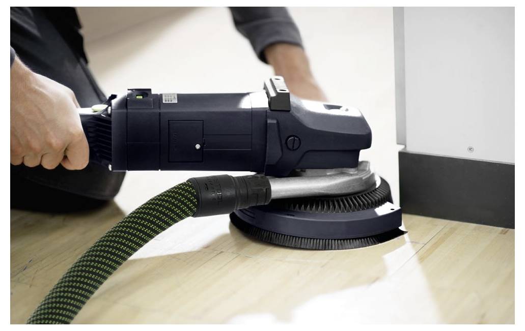 Osoba koristi elektricnu brusiljicu za poliranje drvenoga poda blizu zida, fokusirajući se na uklanjanje nepravilnosti i postizanje glatke površine.