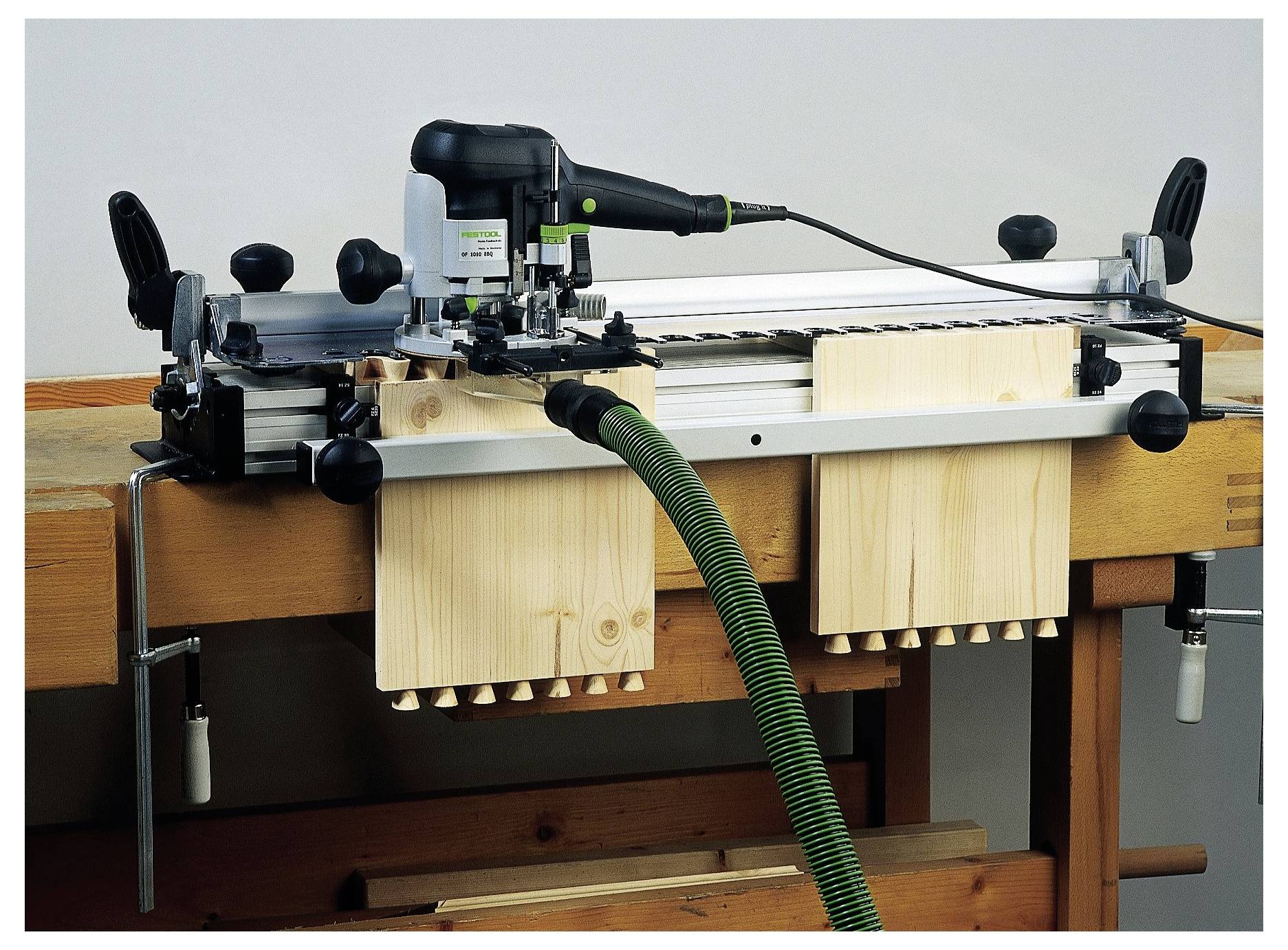 'Tesarski šablon za lastavičaste spojeve na radnoj klupici, korišten za precizno rezanje spojeva. Opremljen je s elektroalatkm i zelenim usisavačem.'