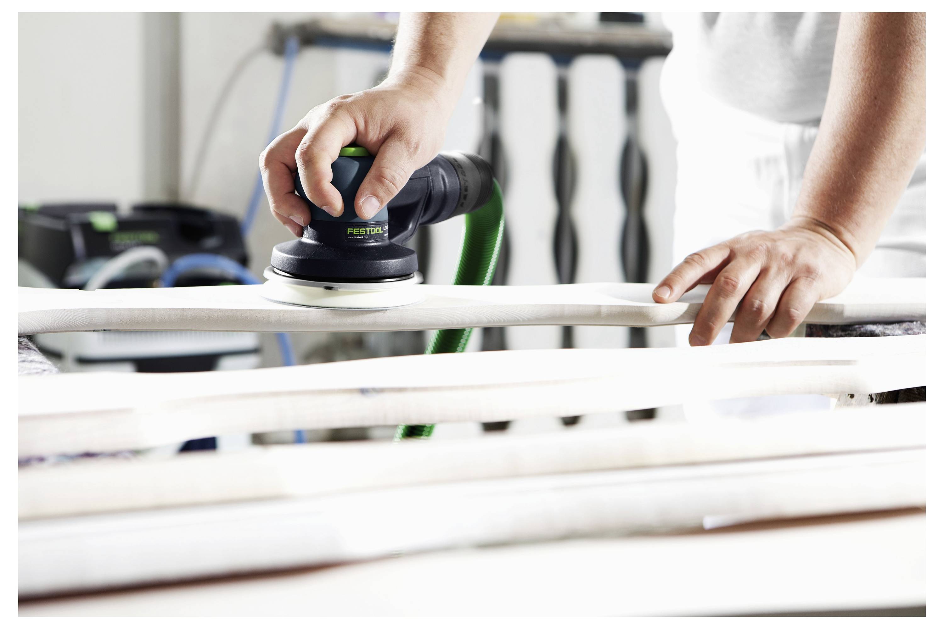 Ruke osobe koja koristi elektricni brusilica za glačanje drvene površine u radionici. U pozadini su vidljivi različiti alati i oprema.