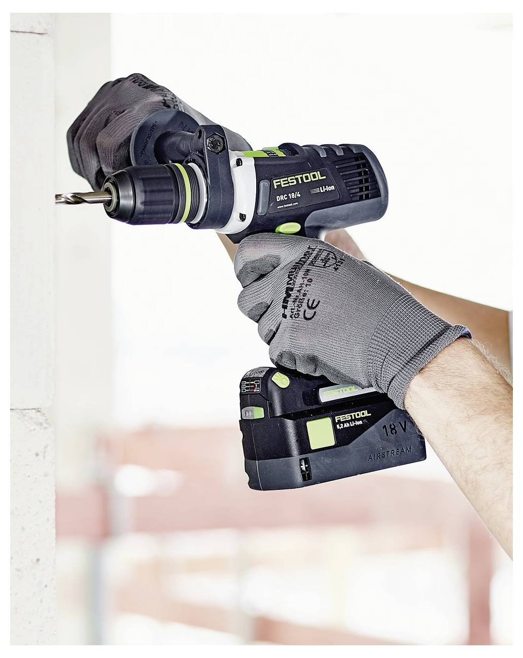 Osoba u rukavicama koristi Festool bušilicu kako bi izbušila rupu u bijelom zidu, što ukazuje na građevinske ili popravne radove.