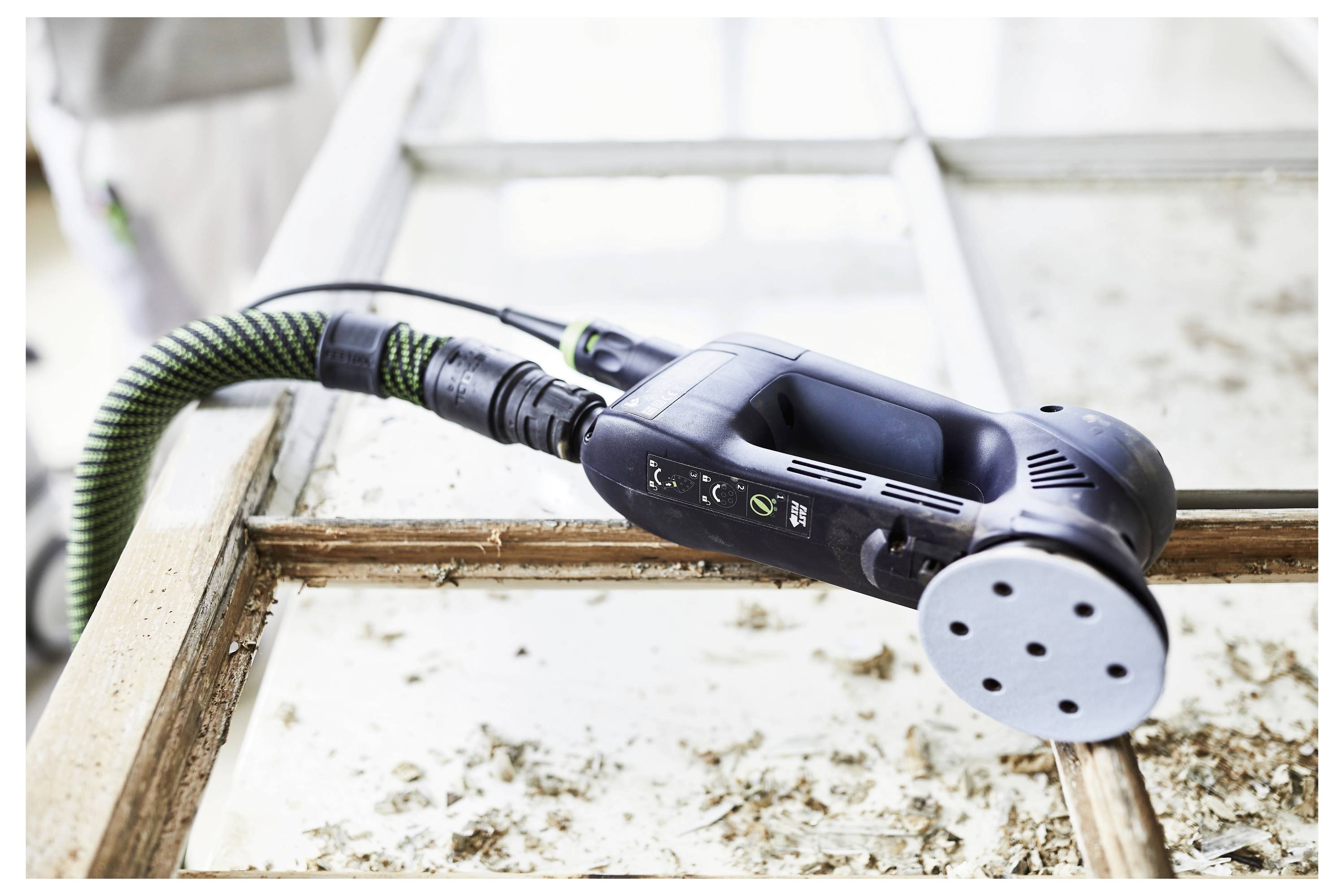 Orbitna brusilica leži na staroj drvenoj prozorskoj rami s ljuštenjem boje i nanosima, što ukazuje na obnovljeni rad u tijeku.