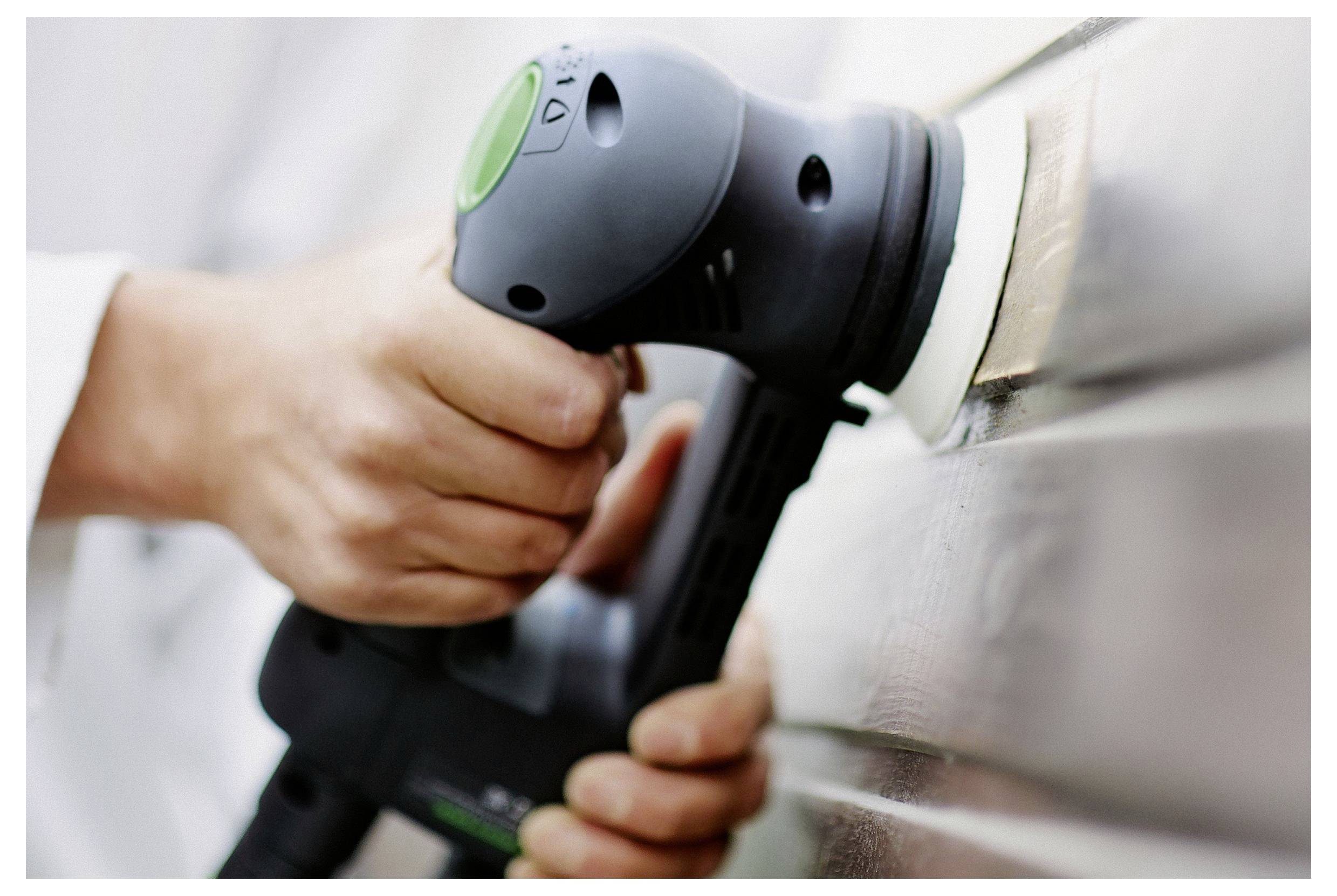 Osoba koristi električni brusilac na drvnoj površini, držeći ga objema rukama. Brusilac je crne boje s zelenim gumbom.