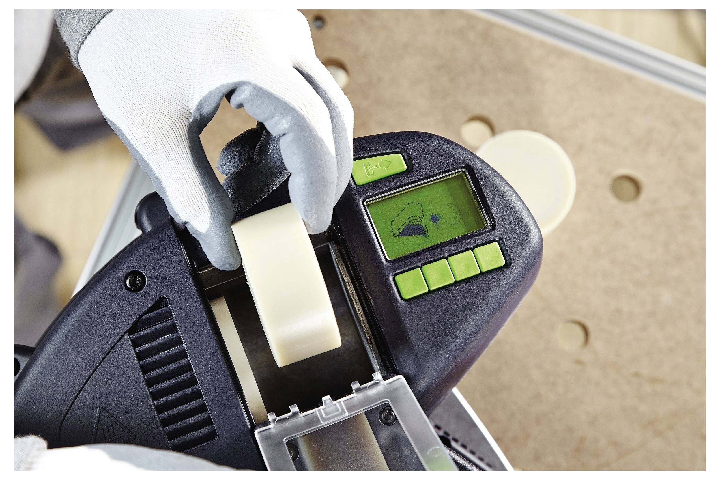 'Osoba koja koristi stroj za nanošenje trake, noseći zaštitne rukavice. Zaslon prikazuje ikonu postavki, dugmići su zeleni, drži rolu trake.'