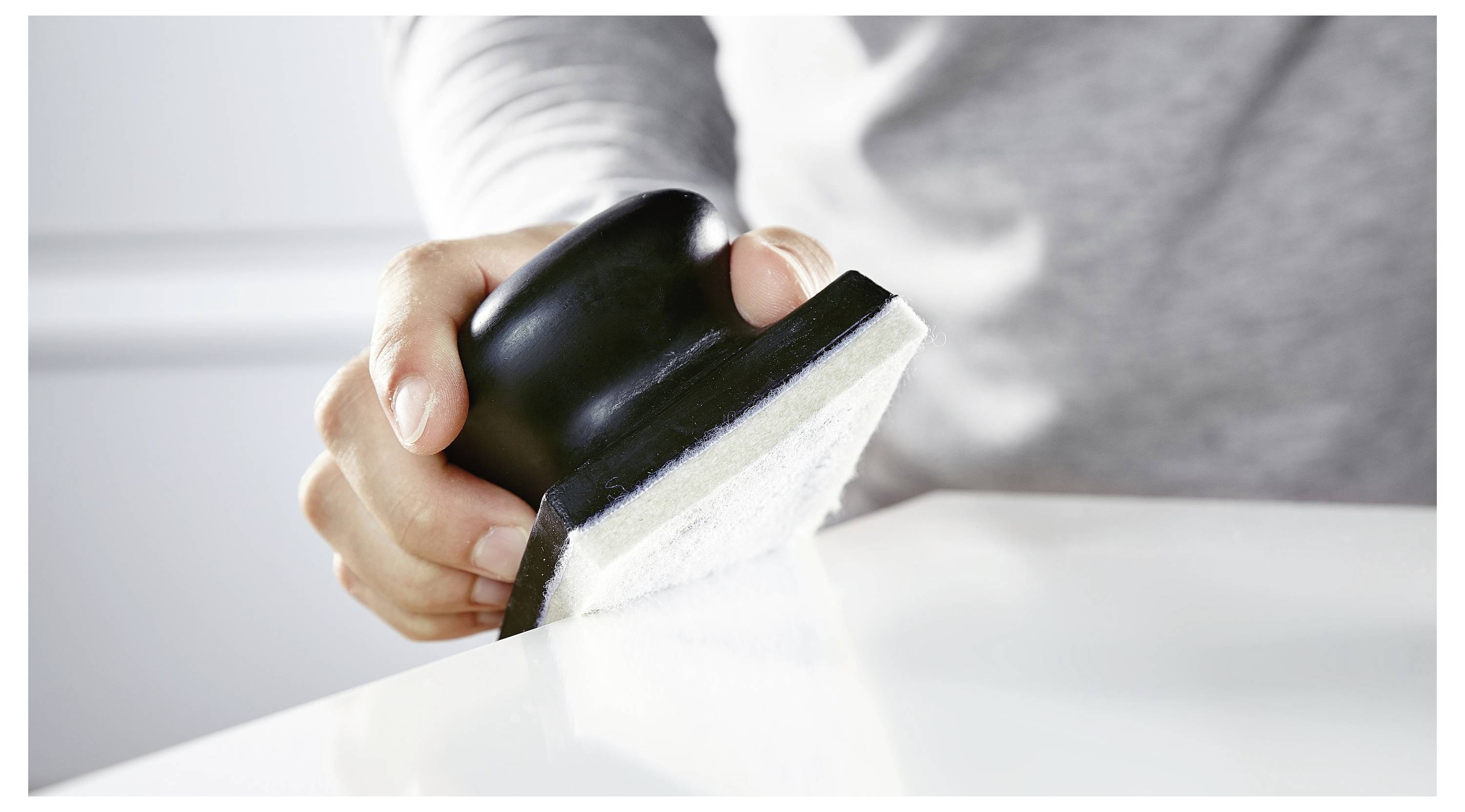 Osoba u sivoj majici koristi alat za čišćenje s crnom ručkom i bijelim jastučićem da obriše glatku, bijelu površinu.