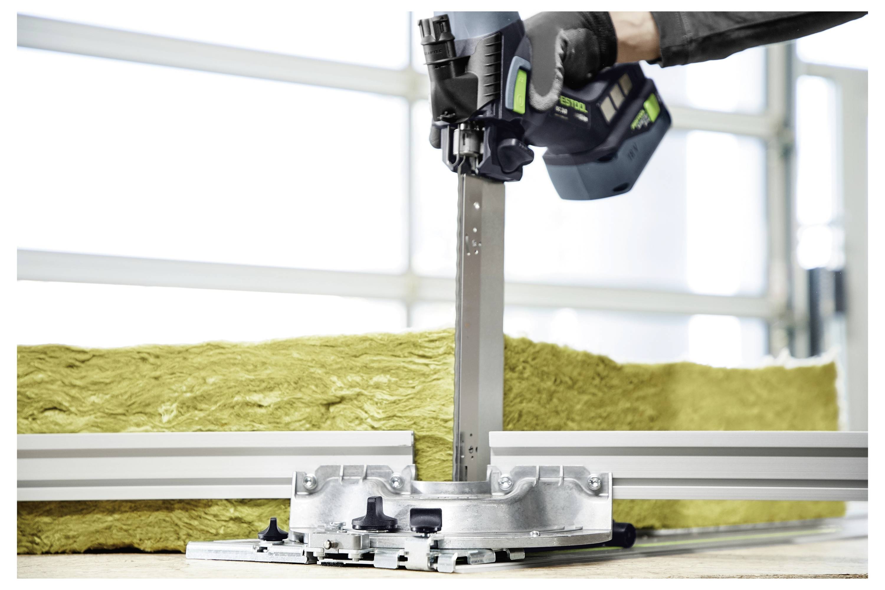 Radnik na gradilištu u bijelom kombinezu koji koristi električni alat za brušenje drvenog dijela kuće. Kutija s alatima nalazi se na skeli.