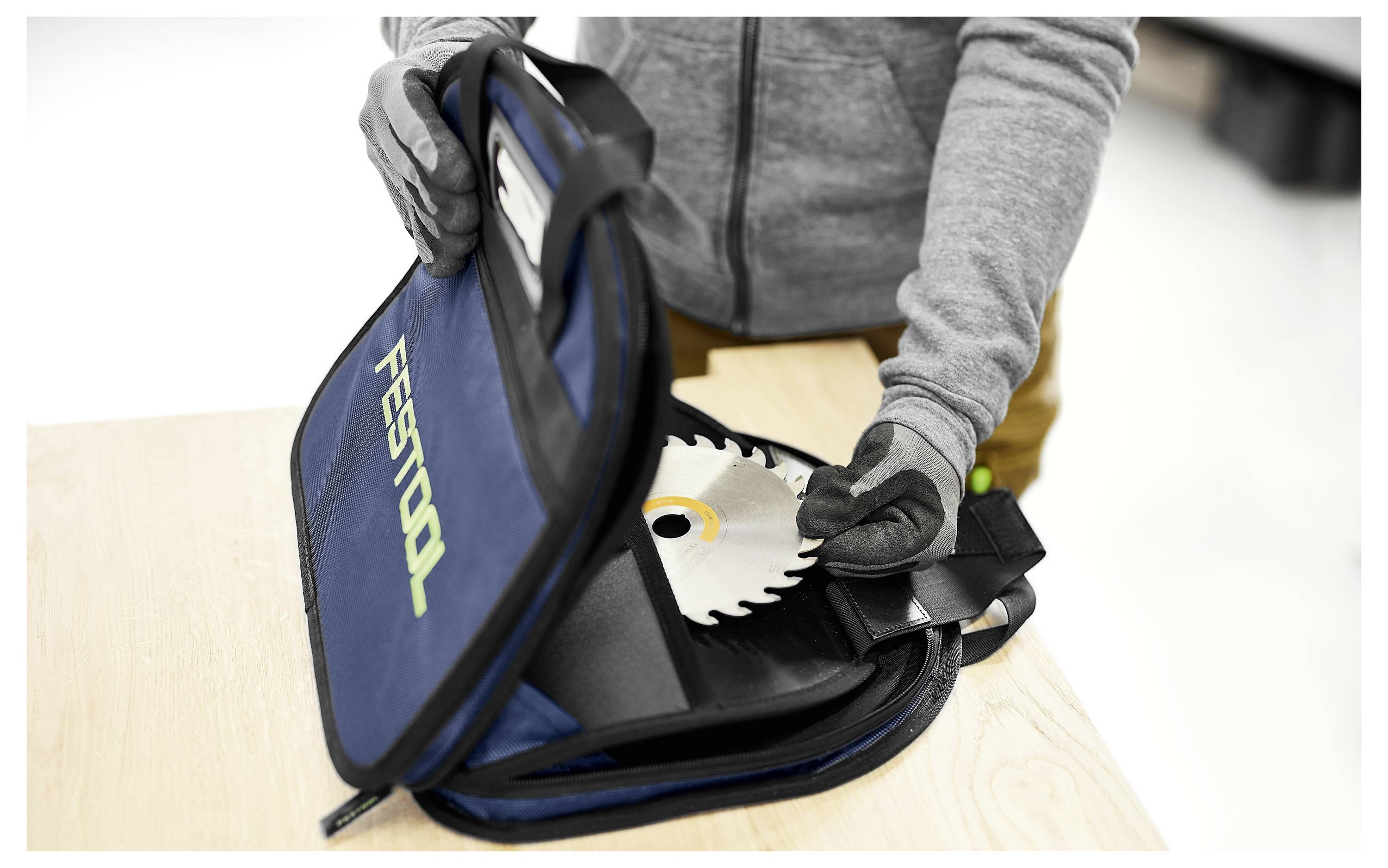 Osoba koja nosi rukavice stavlja žilu kružne pile u plavu Festool torbu na drvenom stolu, što sugerira siguran transport ili skladištenje.
