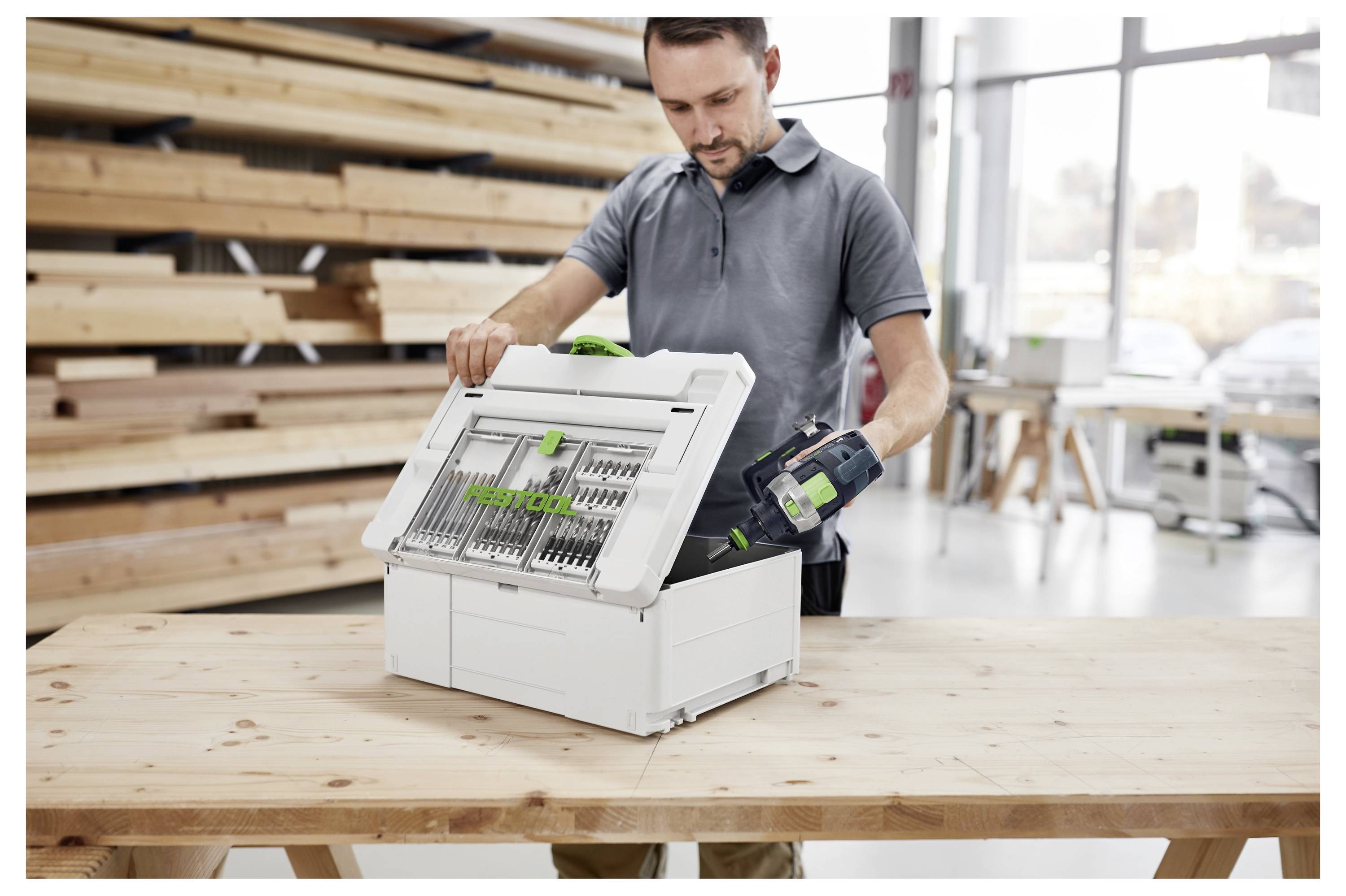 Muškarac koji stoji na ljestvama koristi bušilicu sa zelenim crijevom priključenim, radeći na bijelom zidu unutar moderne industrijske postavke.