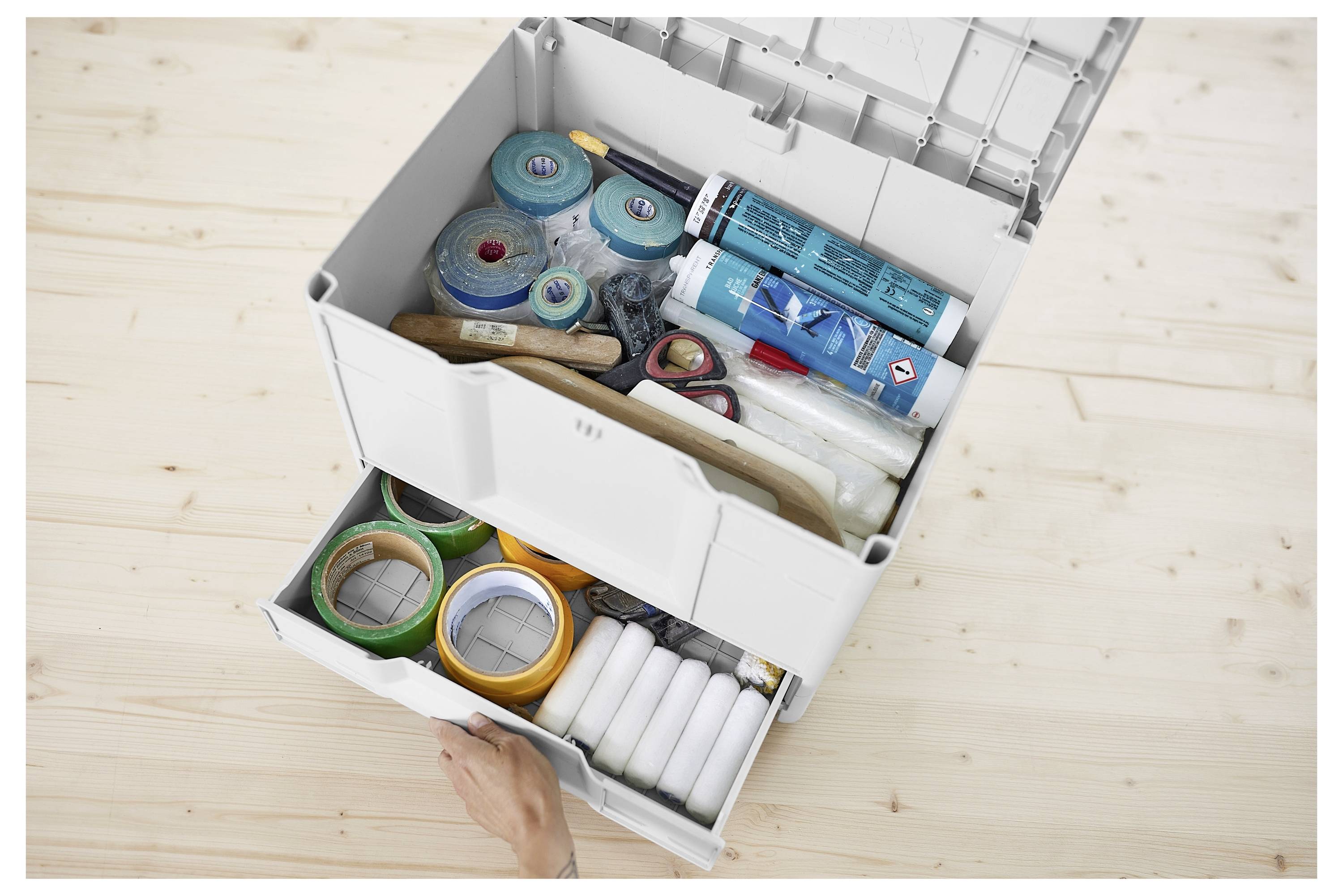 Otvorena kutija za pohranu na drvenom podu koja sadrži različite alate i materijale uključujući role trake, valjke za bojanje, kistove i tube za silikon.