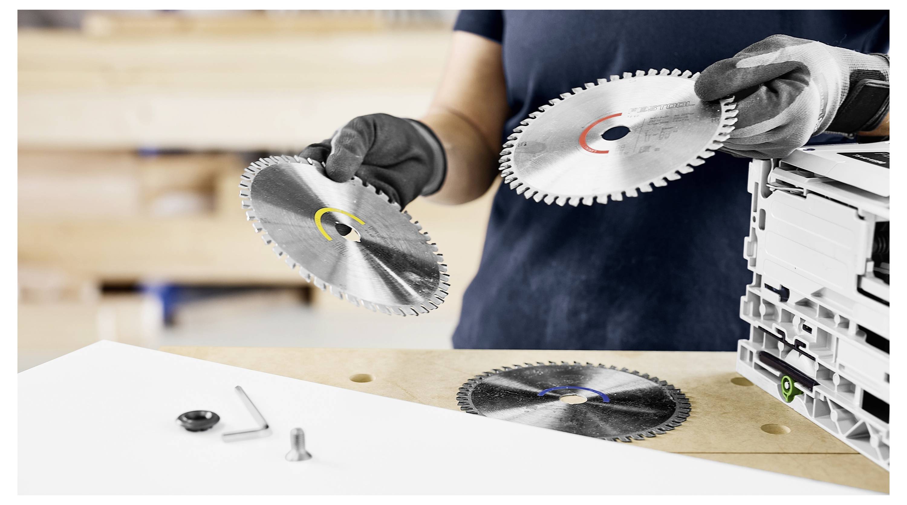 Osoba s rukavicama drži dvije kružne pile, jednu u svakoj ruci, iznad radnog stola. Još jedna pila i nekoliko alata leže na stolu.