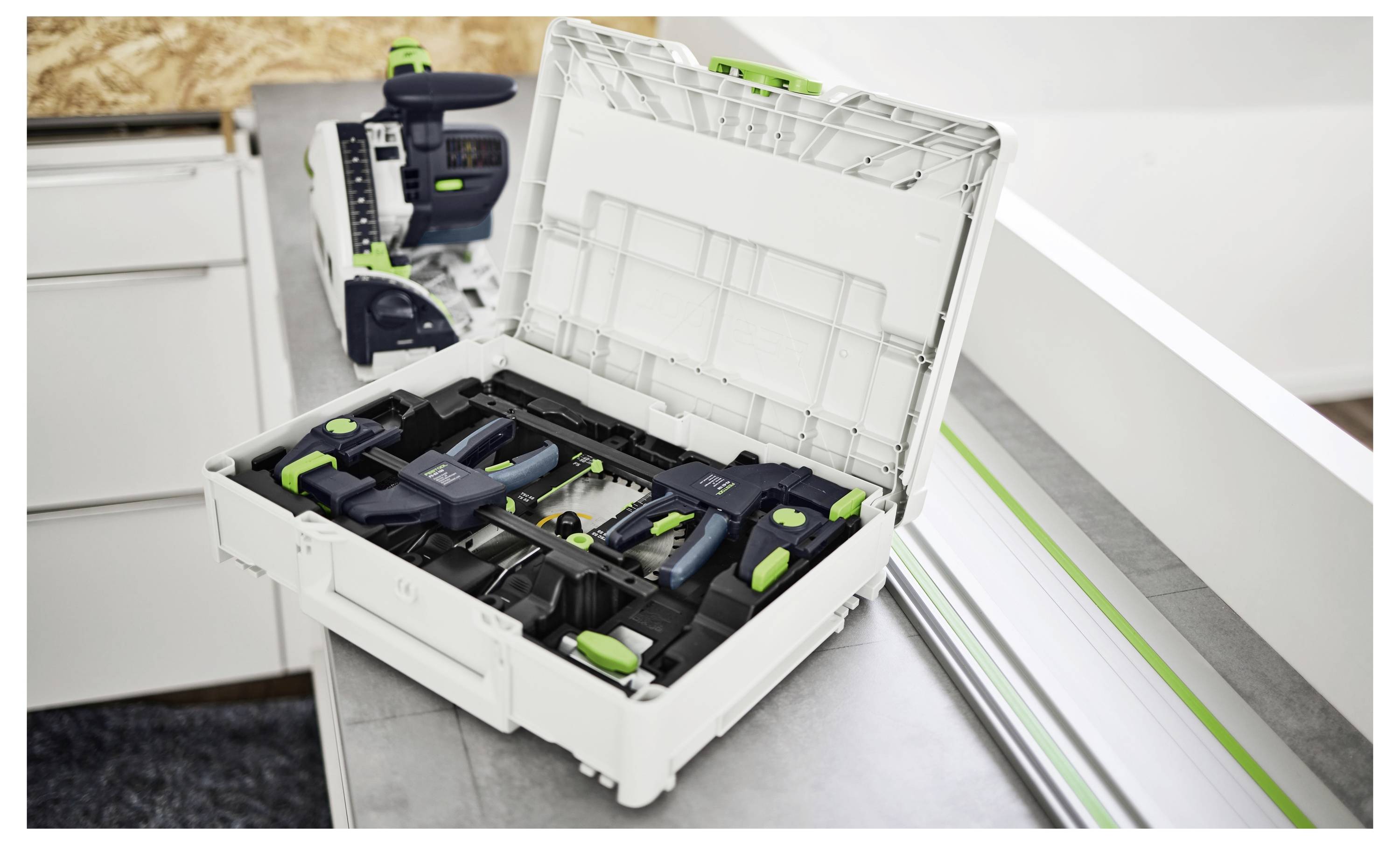 Otvoreni bijeli alat za alat na radnom stolu koji sadrži razne zelene i crne alate s djelomično vidljivim strojem u pozadini.