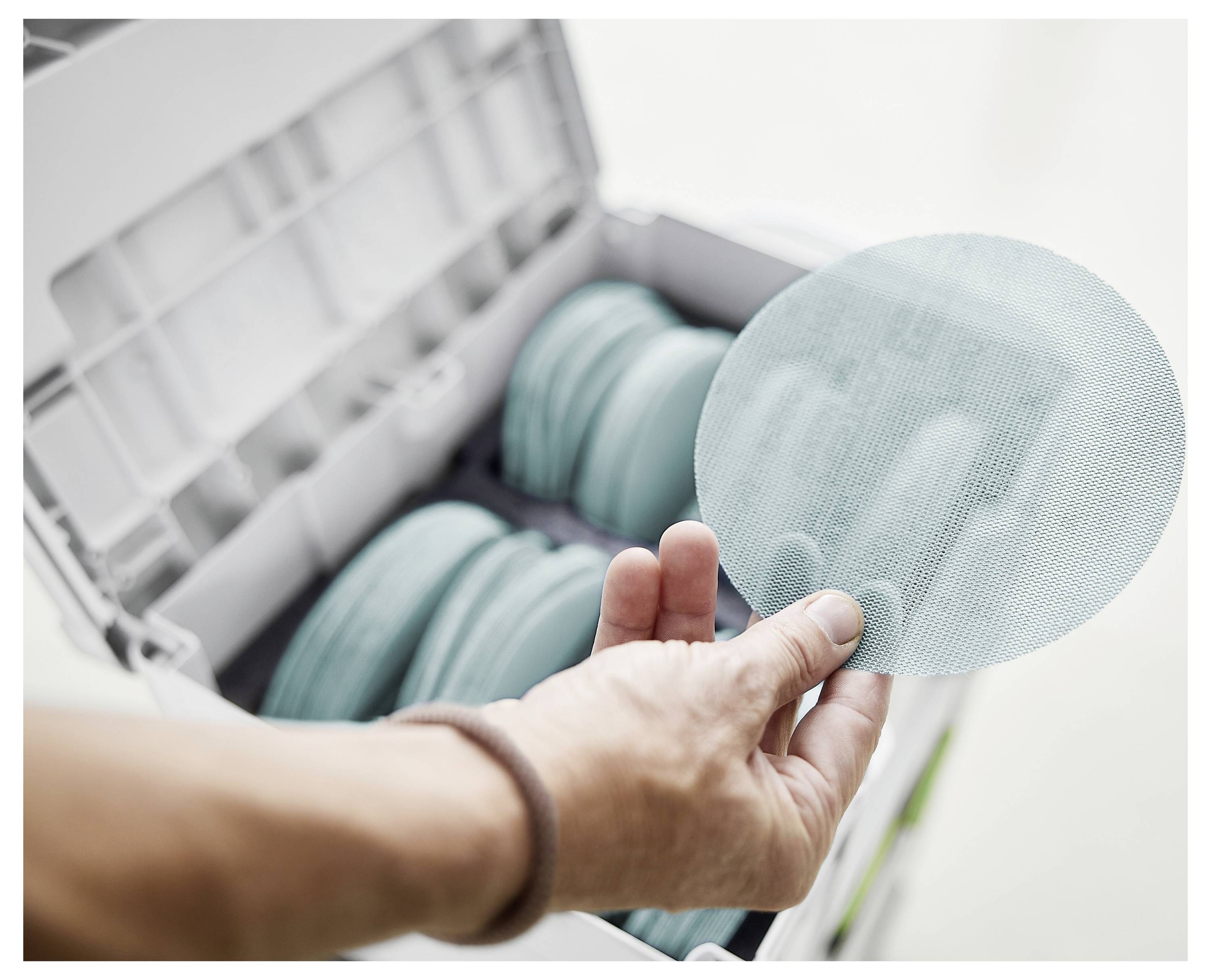 'Osoba drži okrugli brusni disk iznad kutije pune sličnih diskova, ističući alate koji se koriste za brušenje ili pripremu površine.'