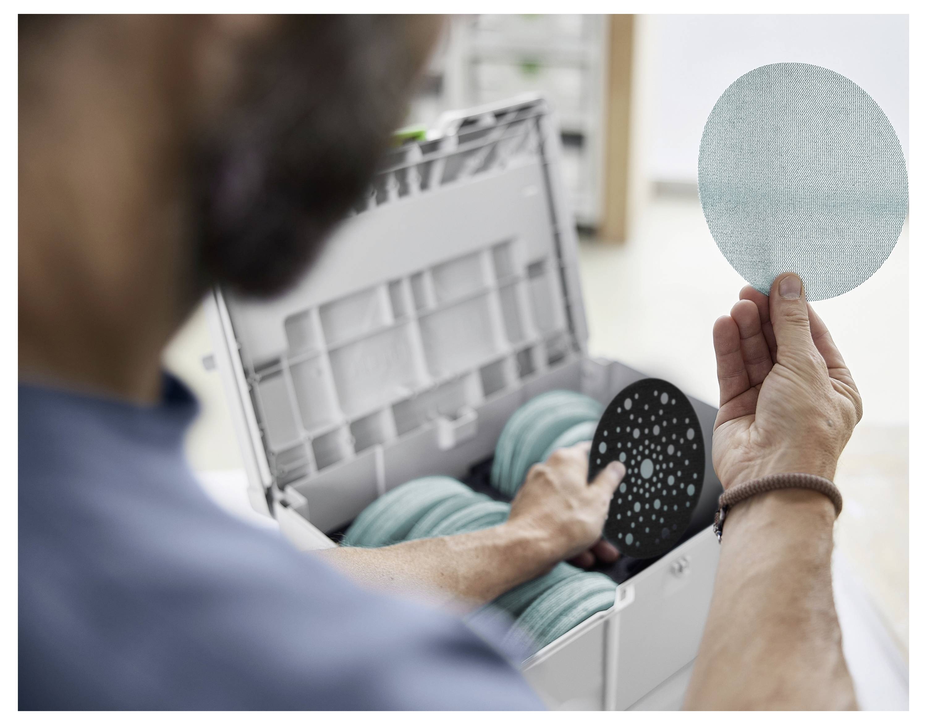 Osoba drži okrugli, svijetloplavi brusni disk u fokusu, s više diskova pohranenih u kutiji. Scena ukazuje na pripreme za brusilice radove.