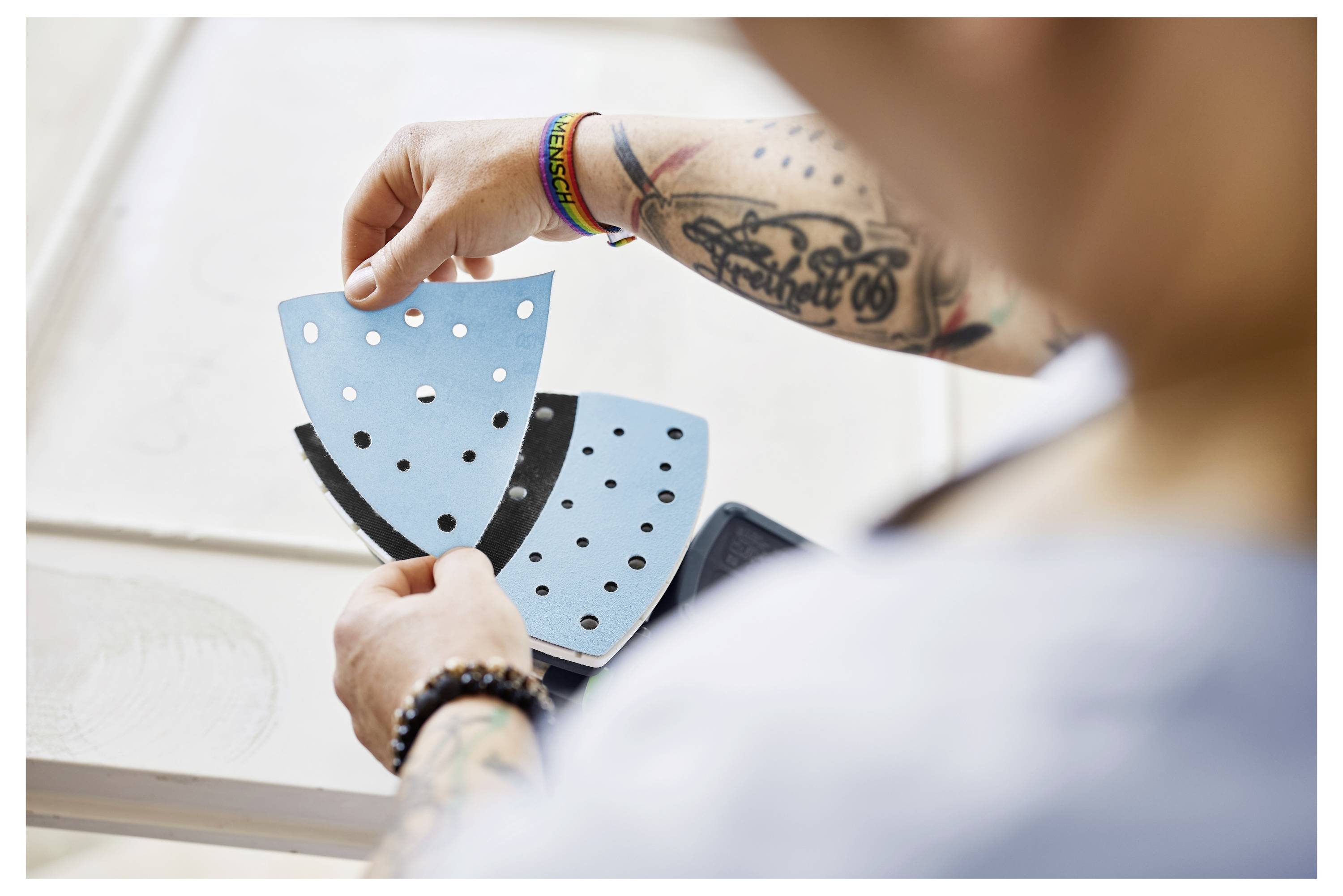 Osoba koja drži plave i crne brusne listove s rupama, noseći narukvicu i imajući vidljive tetovaže, fokusirajući se na materijale.