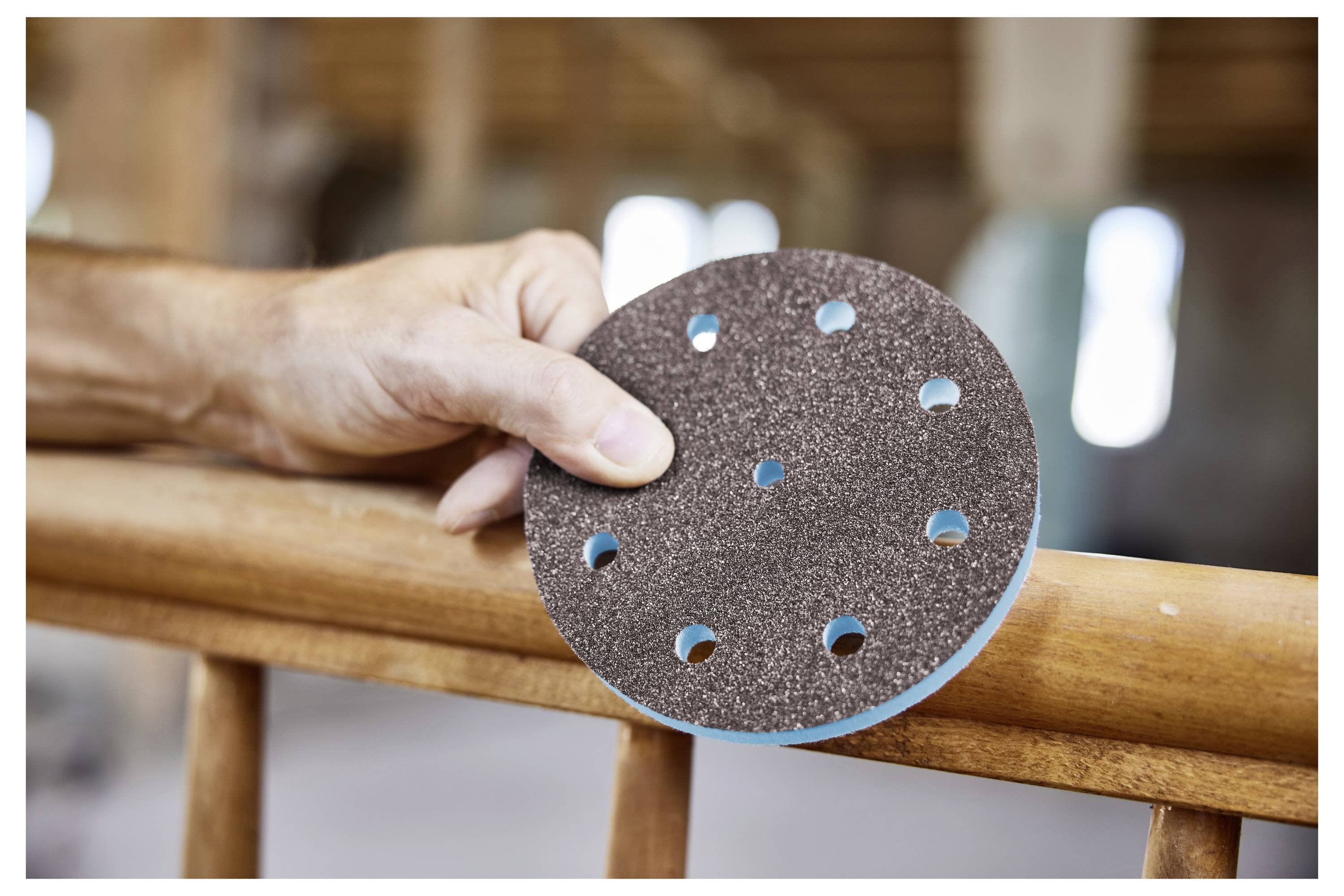 Osoba drži kružni brusni disk rukom pritisnuvši ga na drvenu balustrad, što ukazuje na woodworking ili radove ojačavanja u tijeku.