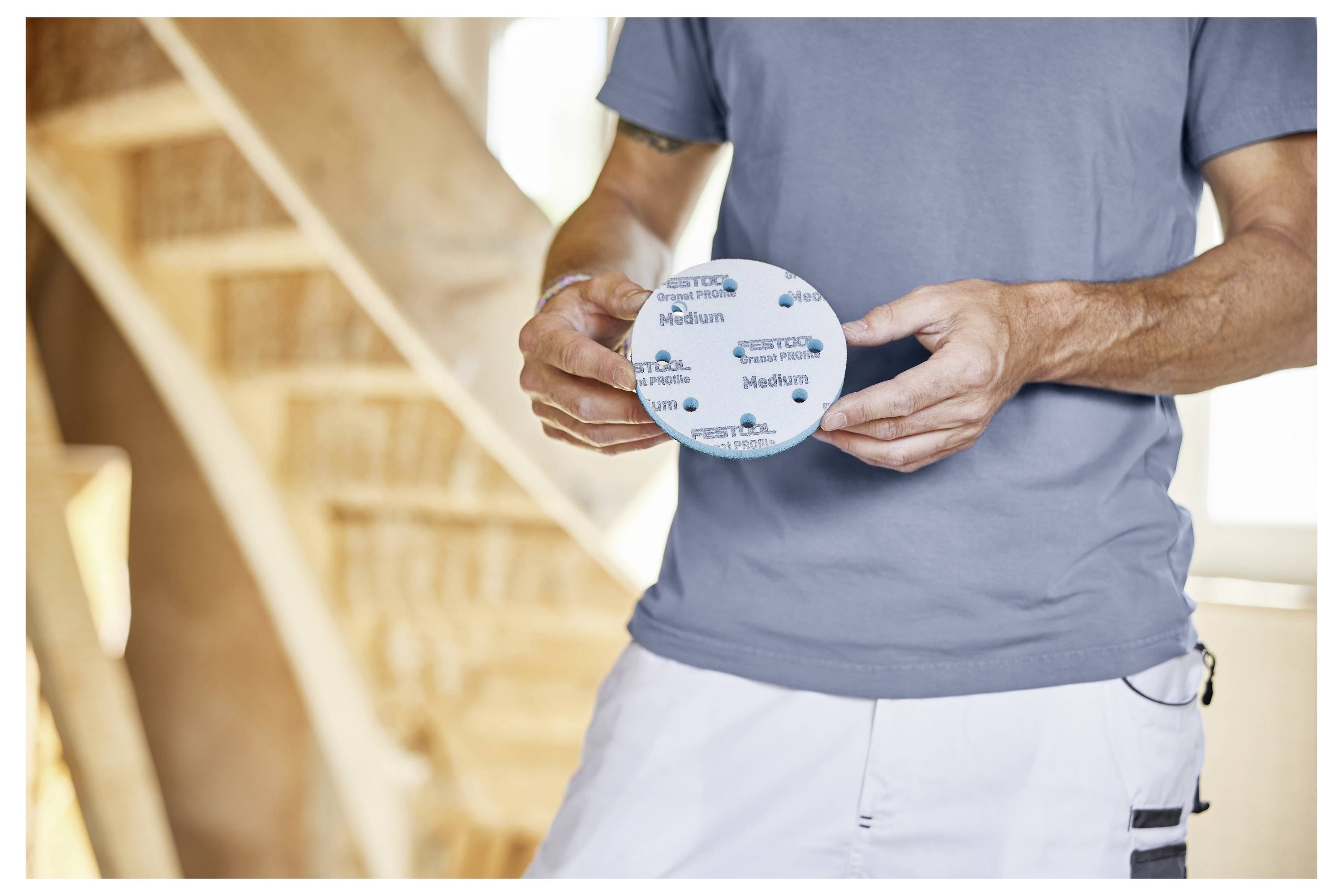 Osoba odjevena u sivi majicu drži okrugli brusni disk s natpisom 'Medium' i 'Festool' blizu drvenih stepenica u izgradnji.