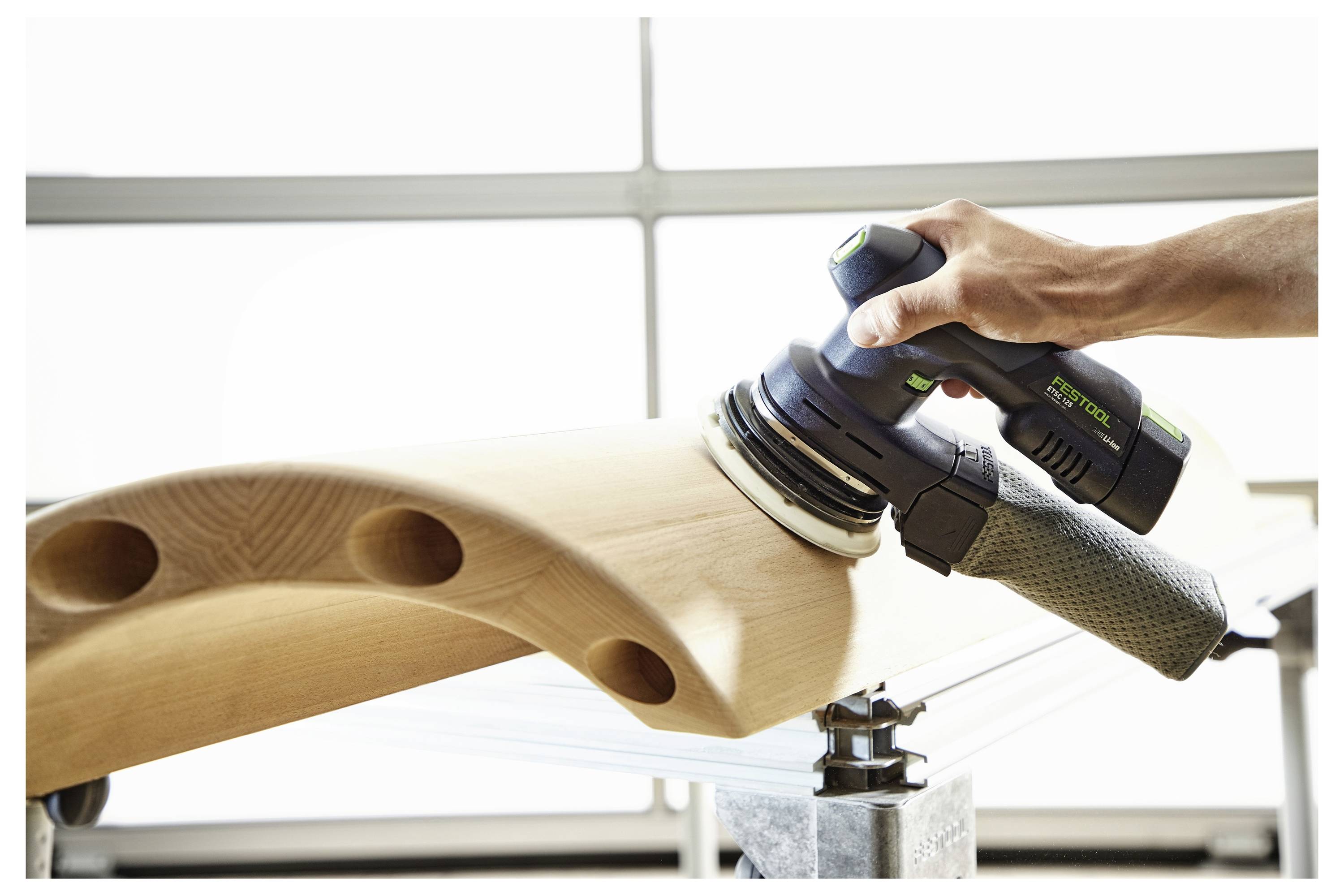 Ruka koja drži blok za brušenje, djelomično umetnut u bijeli džep radnog pojasa, sa zamućenom drvenom strukturom u pozadini.