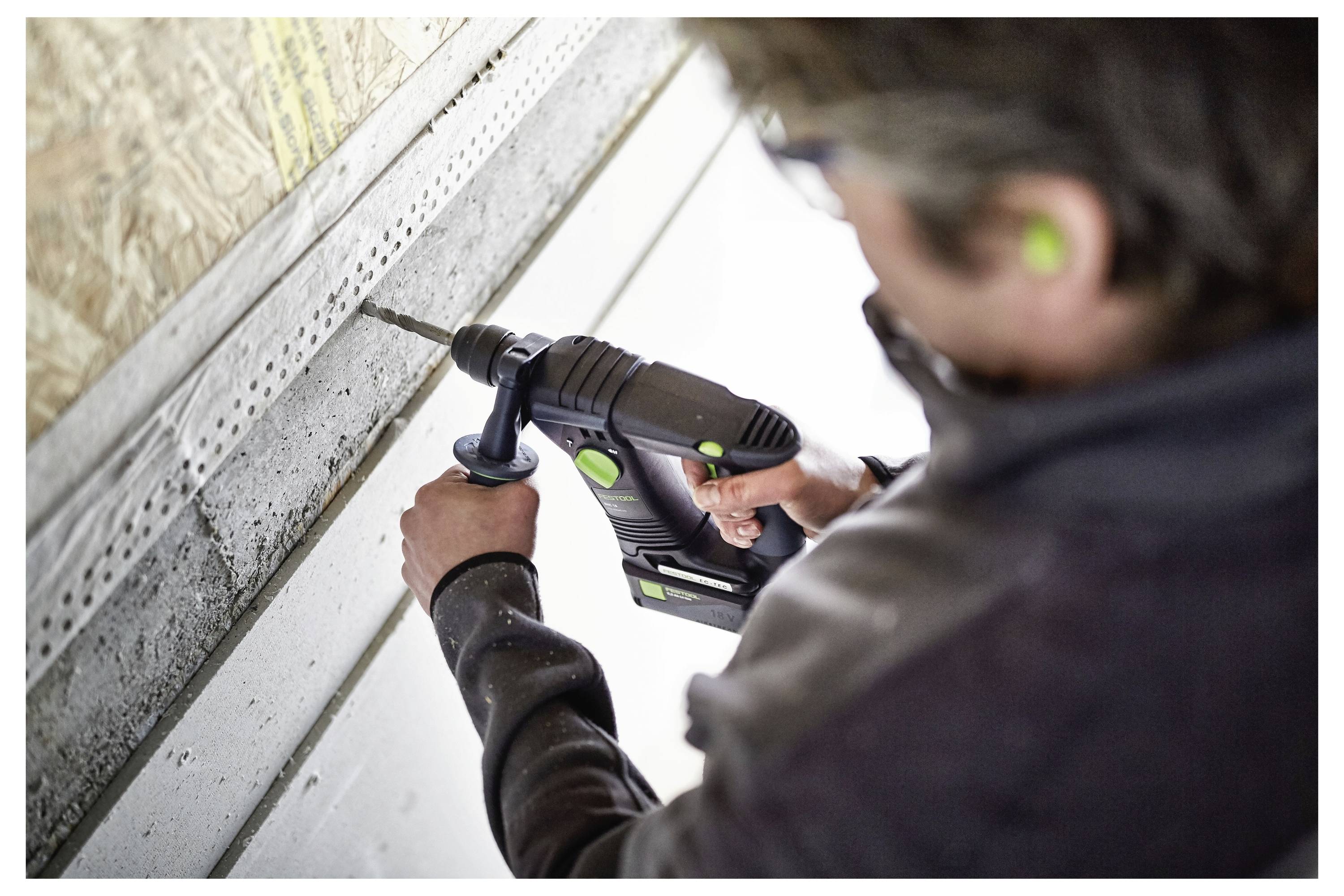 Osoba koristi elektrnu bušilicu na zidu dok nosi zaštitnu opremu kao što su čepići za uši, što sugeriše gradnju ili instalacijskie radove.