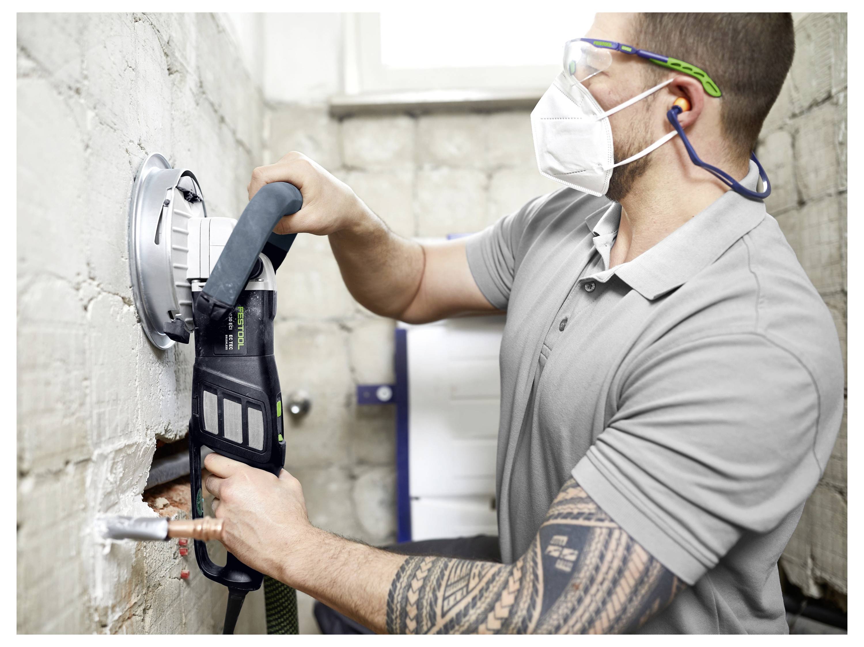 Osoba u zaštitnoj opremi koristi elektroalat za rezanje u bijelu zidnu konstrukciju od opeke, otkrivajući cijevi.