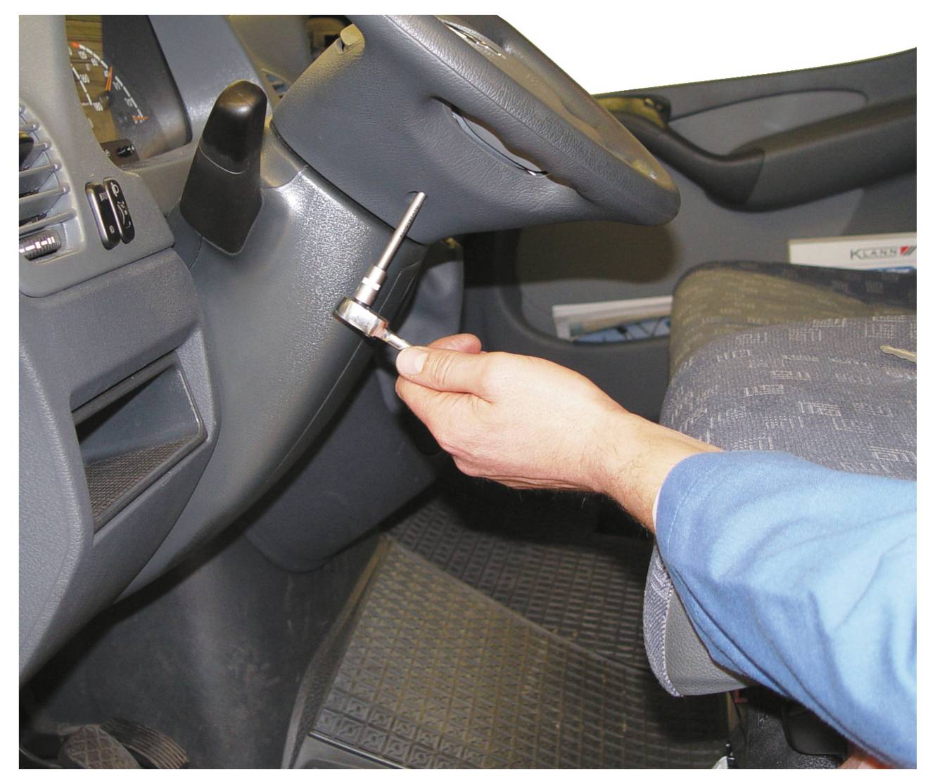 'Osoba koja koristi nastavnu ključ na vijku ispod vozačke ploče; vidljiva plava rukava; prikazan unutrašnjost vozila i volan.'