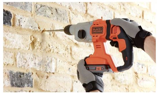 Osoba u rukavicama koristi narančasto-crnu električnu bušilicu kako bi izbušila rupu u zidu od cigle.