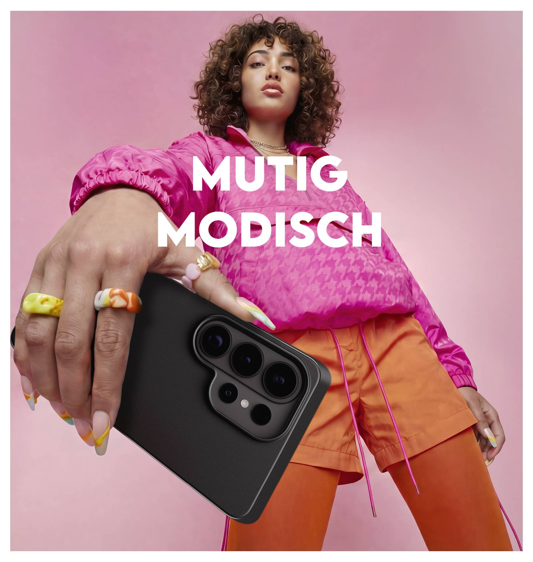 Osoba u ružičastoj jakni i narančastim šorcima drži pametni telefon prema kameri. Tekst glasi „Mutig Modisch", što znači „Hrabro Moderno".