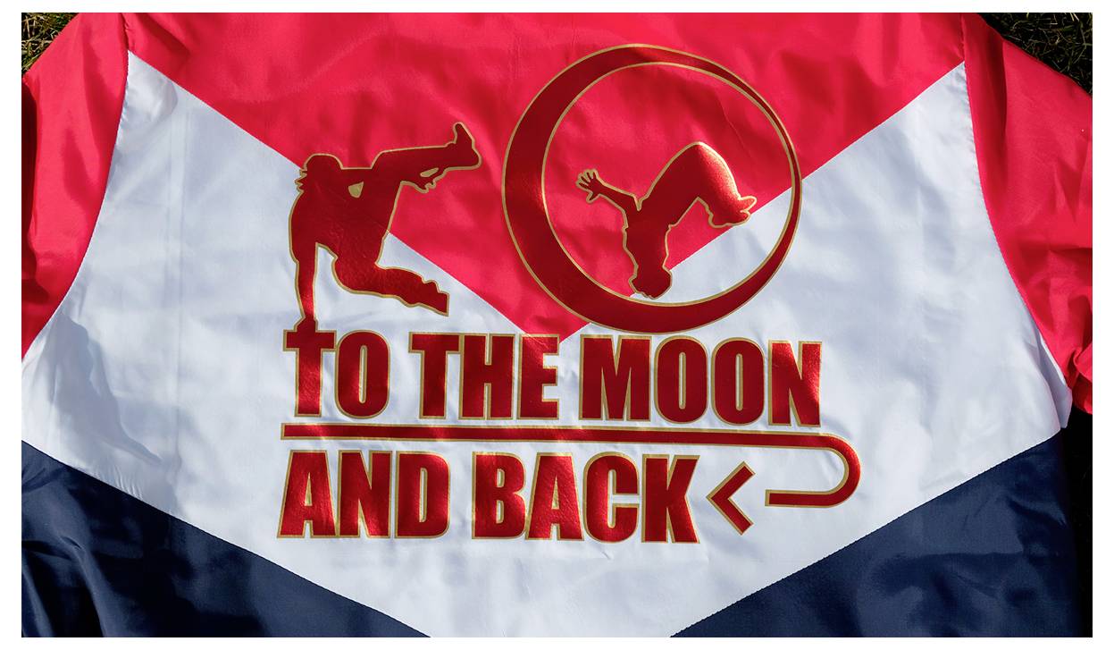 Crvenoj, bijeloj i plavoj boji dizajn s sjenicama dva akrobata i debelog teksta 'TO THE MOON AND BACK' s strelicom ispod.