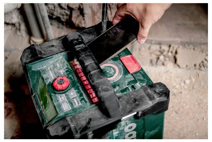 Osoba u radnoj odjeći drži kovčeg alata ''Metabo'', stojeći u dijelom završenoj sobi s izloženim drvenim gredama i radnom svjetiljkom.