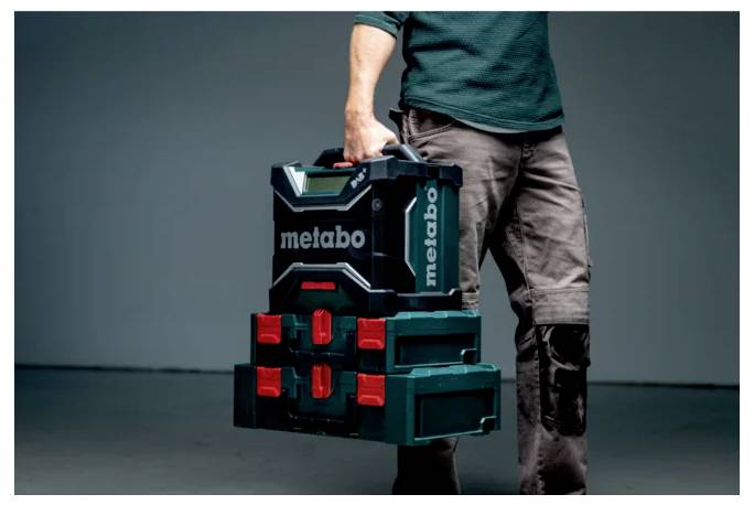 Osoba koja nosi slaganu Metabo kutiju za alate s ručkom, odjevena u radne hlače, pokazujući prenosivost i lakoću transporta.