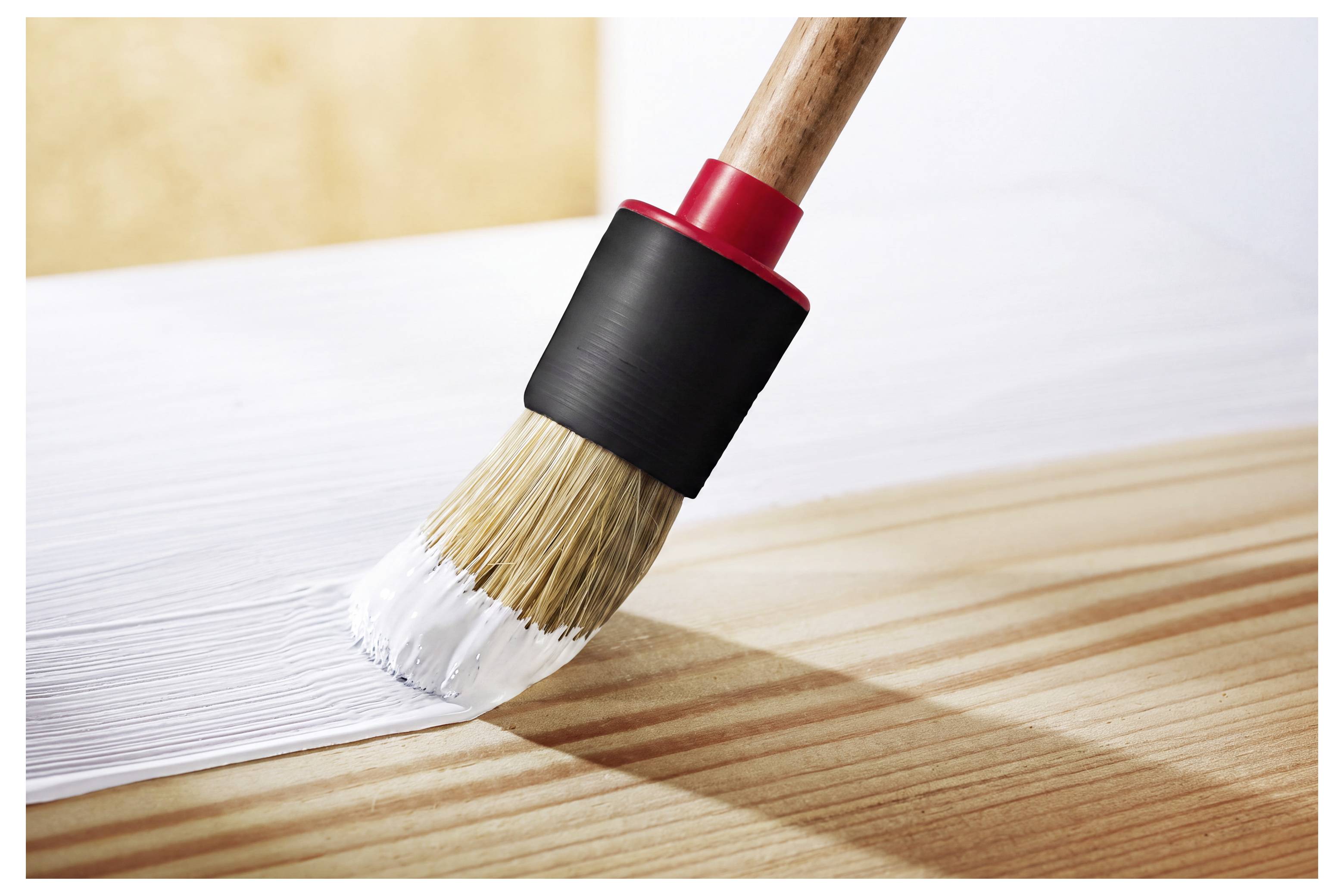 Četka za slikanje koja nanosi bijelu boju na drvenu površinu, ostavljajući svježi sloj i vidljiv potez četke.