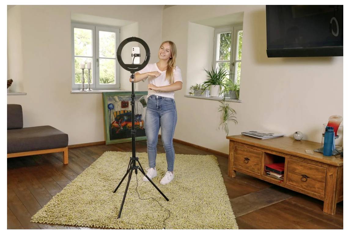 Mlada žena podešava kružno svjetlo na trepezdu u svjetloj dnevnoj sobi sa sofom, tepihom, TV-om i drvenom ormarom.