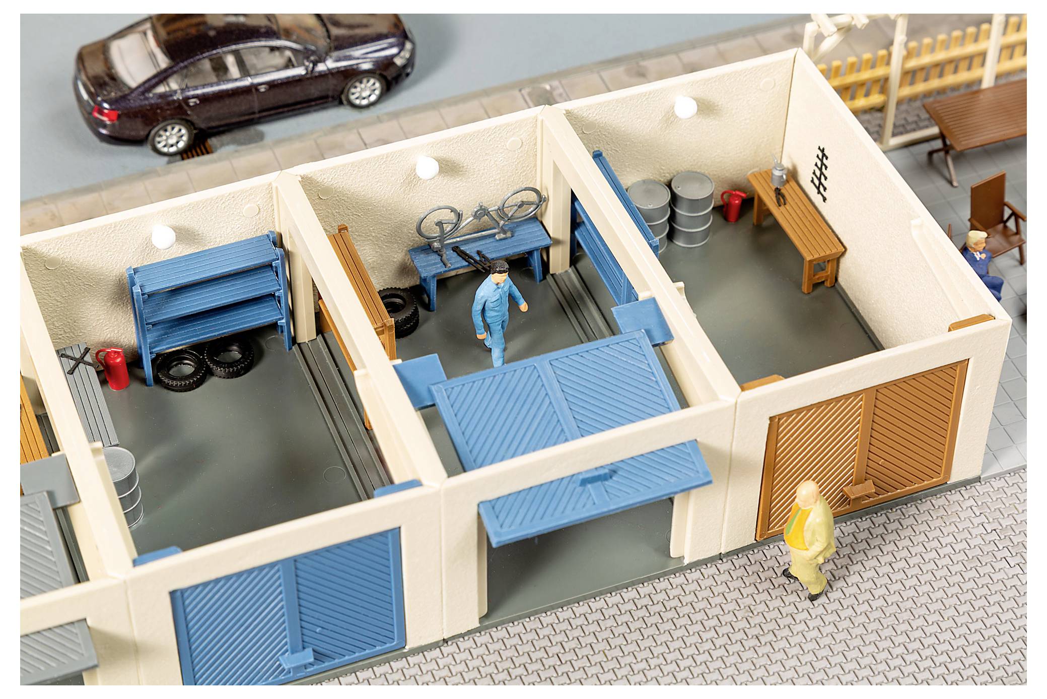 Skladišni/radioničarski diorama igračke s pogledom iz ptičje perspektive s plavim vratima garaže, radnicima, naslaganih bačvi, biciklom i parkiranih automobila vani.