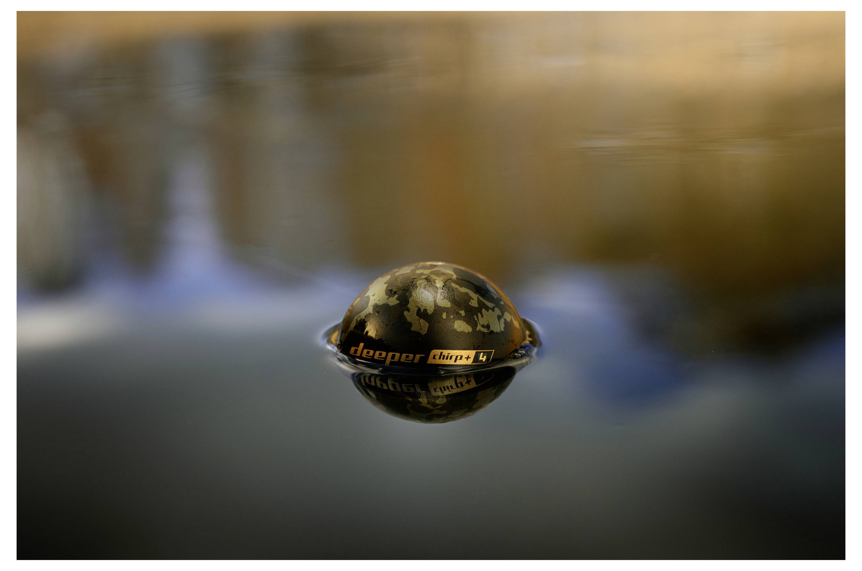 Kamuflirana Deeper sonar naprava pluta na mirnoj vodi, reflektujući se ispod zamućene, toplih tonova obale.