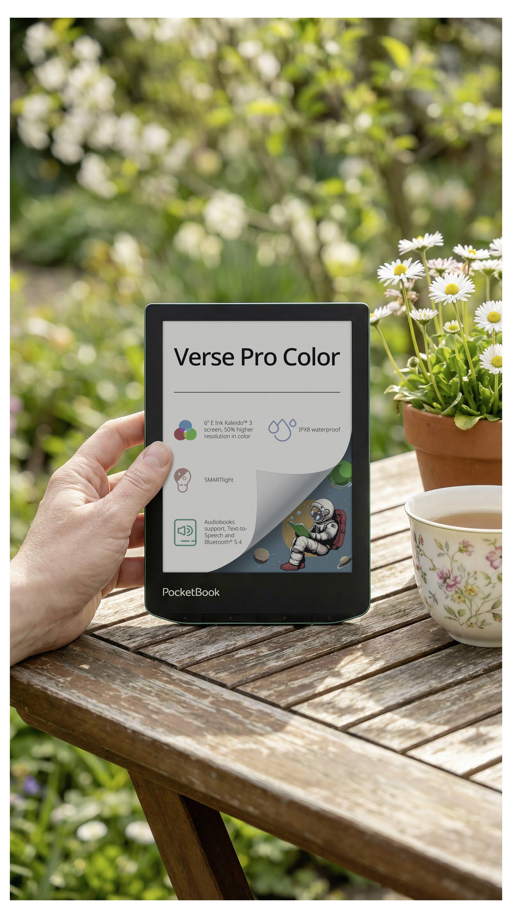 Ruka koja drži PocketBook e-čitač na vrtnom stolu; zaslon prikazuje 'Verse Pro Color' s promocijom proizvoda, pored tratinčica i šalice čaja.